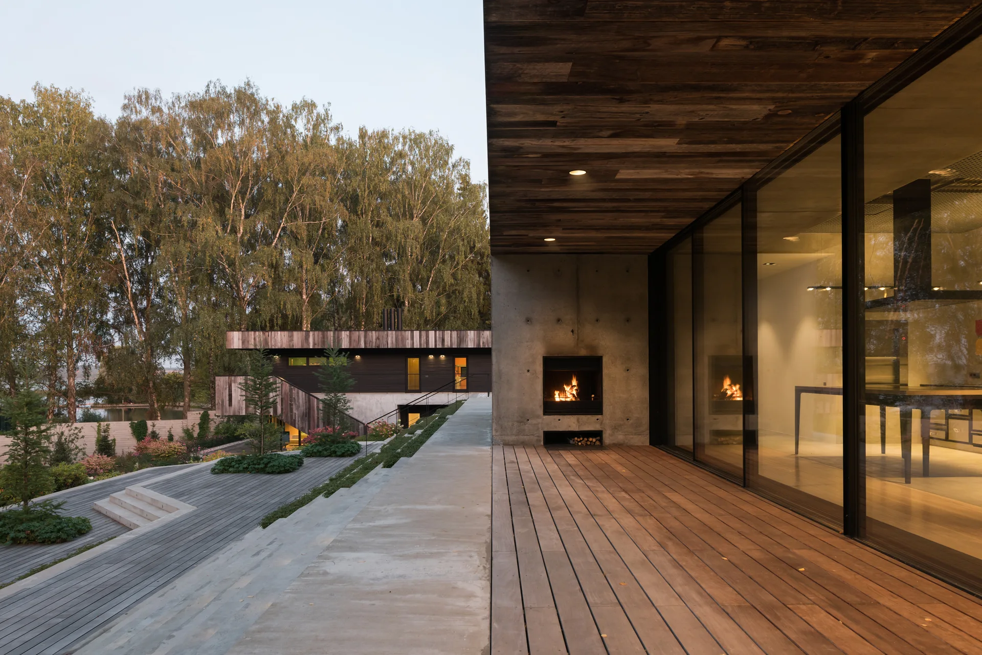 A covered wooden patio features a raw concrete wall with a burning fireplace next to large glass doors looking out onto a terraced garden.