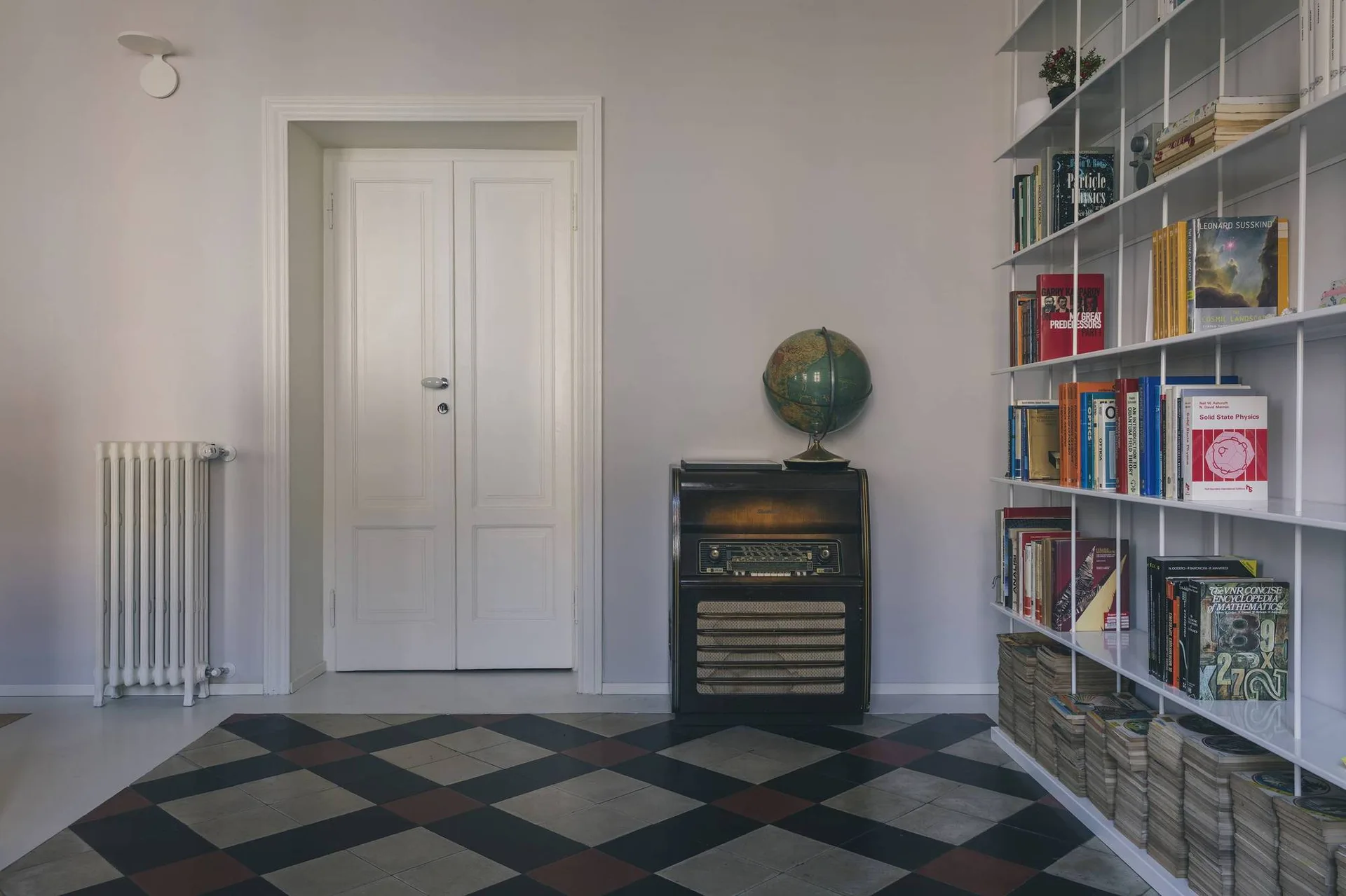 An interior space features a diamond-patterned tile floor, white double doors, a vintage radio topped with a globe, and a white metal bookshelf.