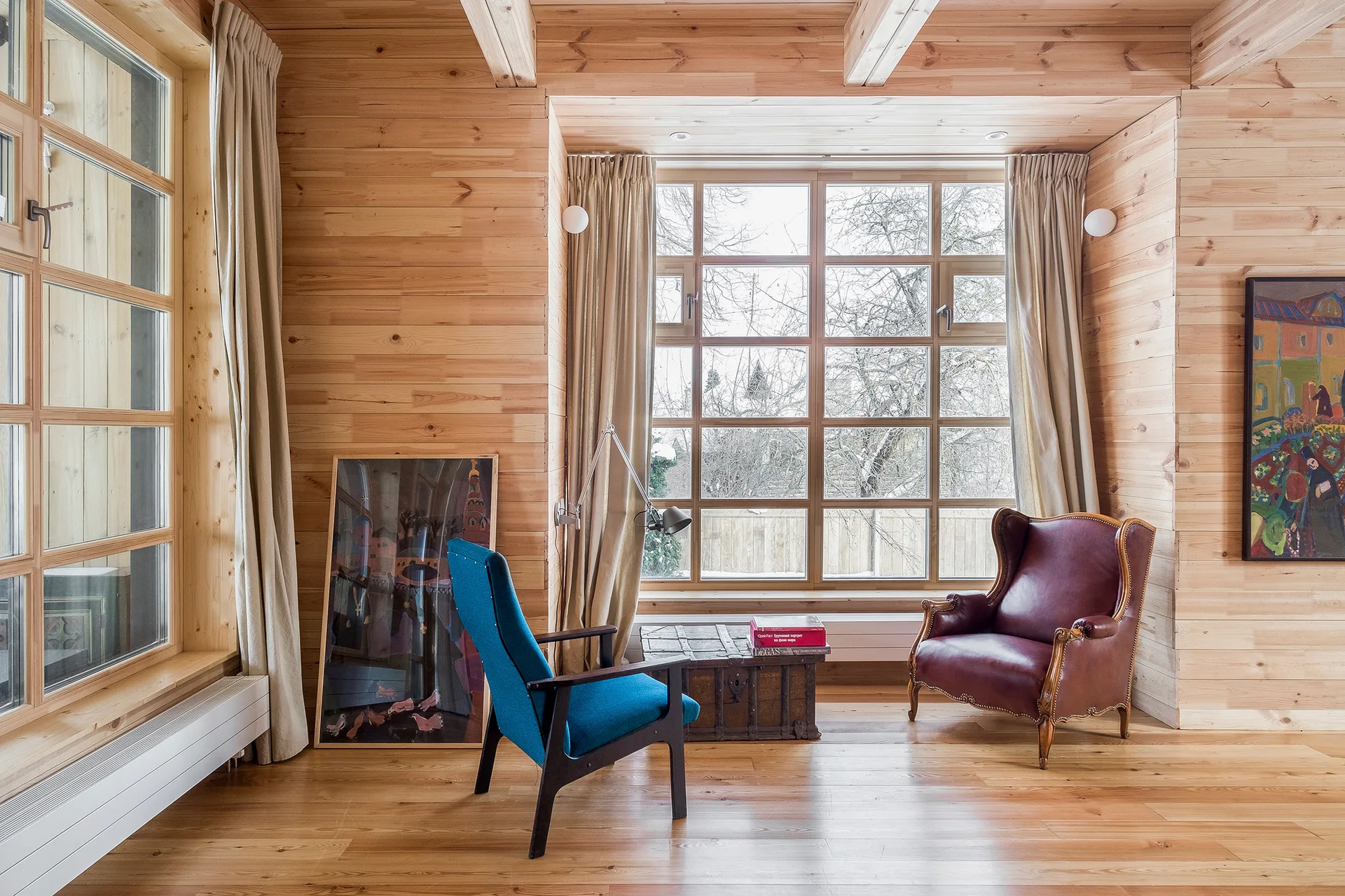A rustic sitting room with horizontal wood paneling features a blue armchair and a burgundy leather wingback chair near a large window.