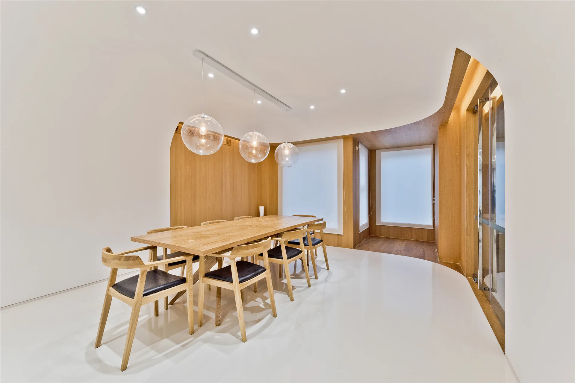 A minimalist dining room featuring a long wooden dining table, black-cushioned chairs, clear glass globe pendant lights, and curved wood paneling.