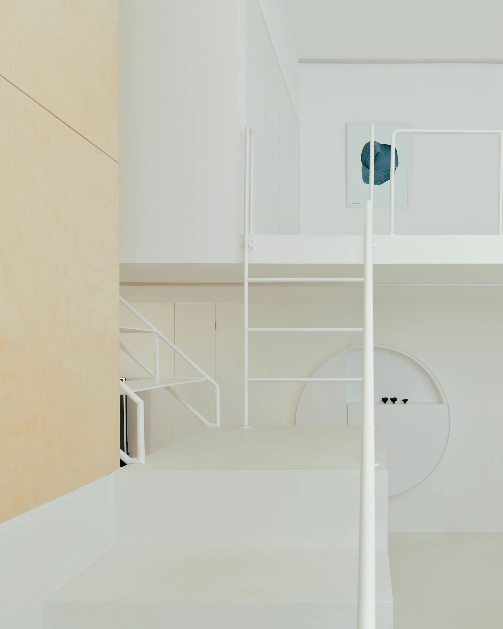A minimalist white loft apartment features a white metal staircase and ladder next to a large light wood plywood wall panel and a circular wall niche.