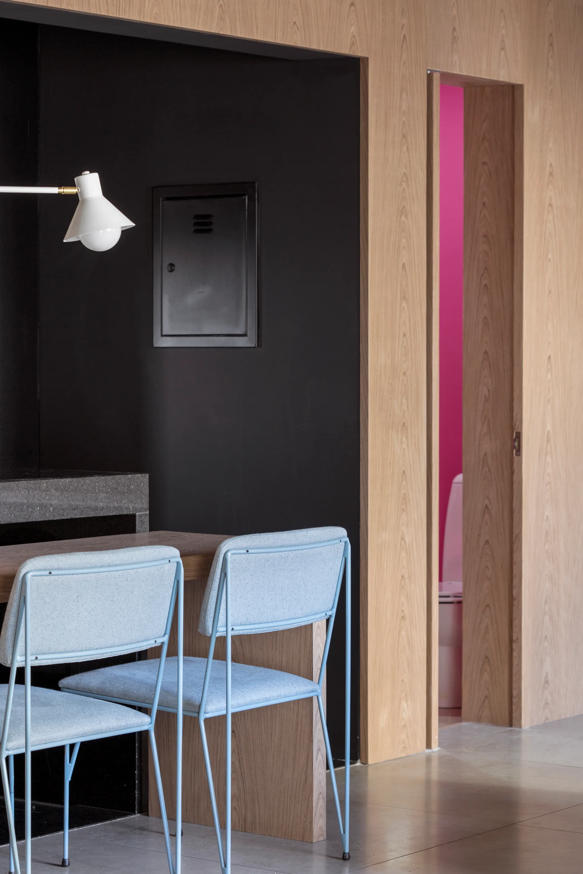 Two light blue chairs are set at a wooden table next to wood-veneered paneling, with a narrow doorway revealing a bright pink bathroom.
