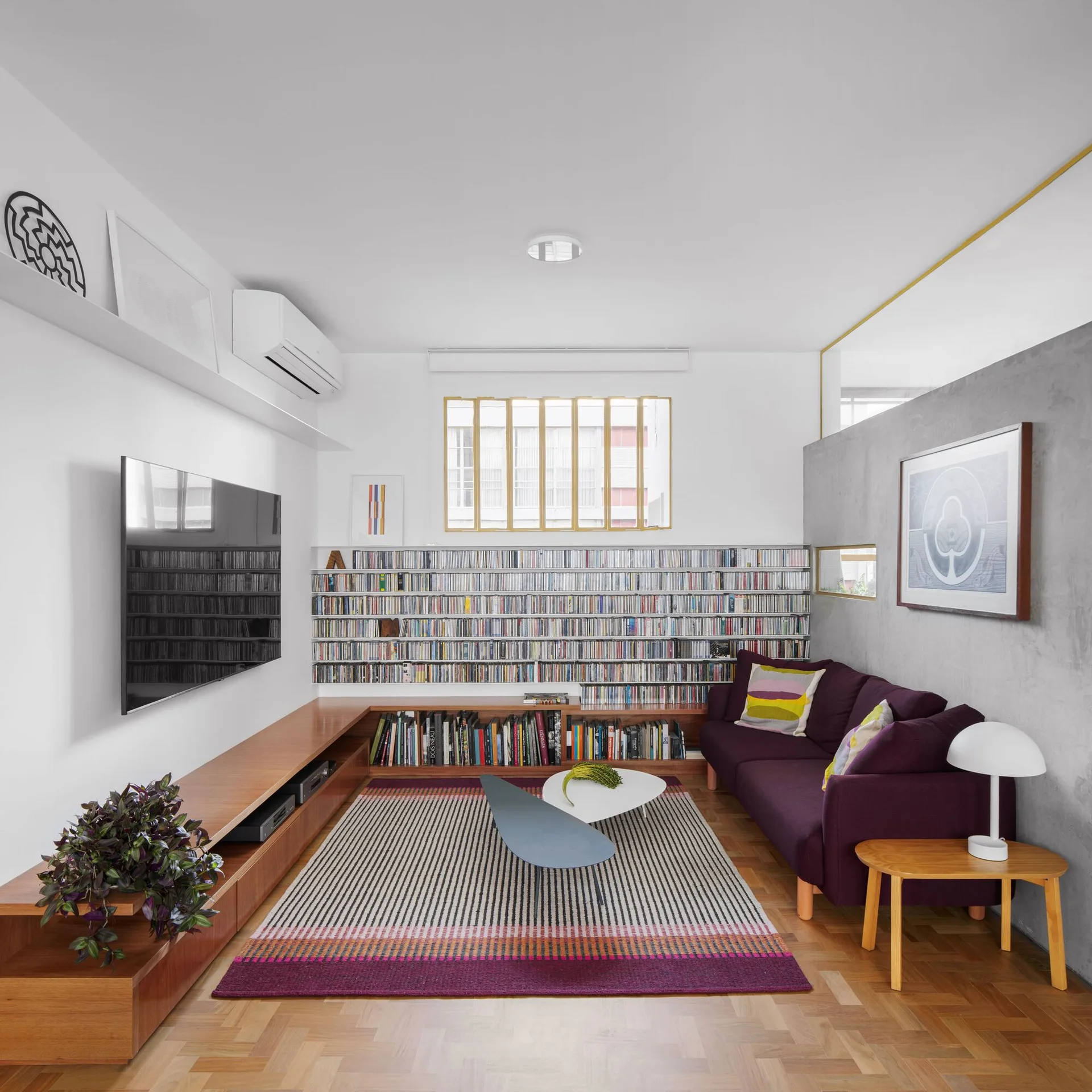 A contemporary living room featuring a purple sofa, a striped area rug, a low wooden media console, and a wall lined with a large CD collection.