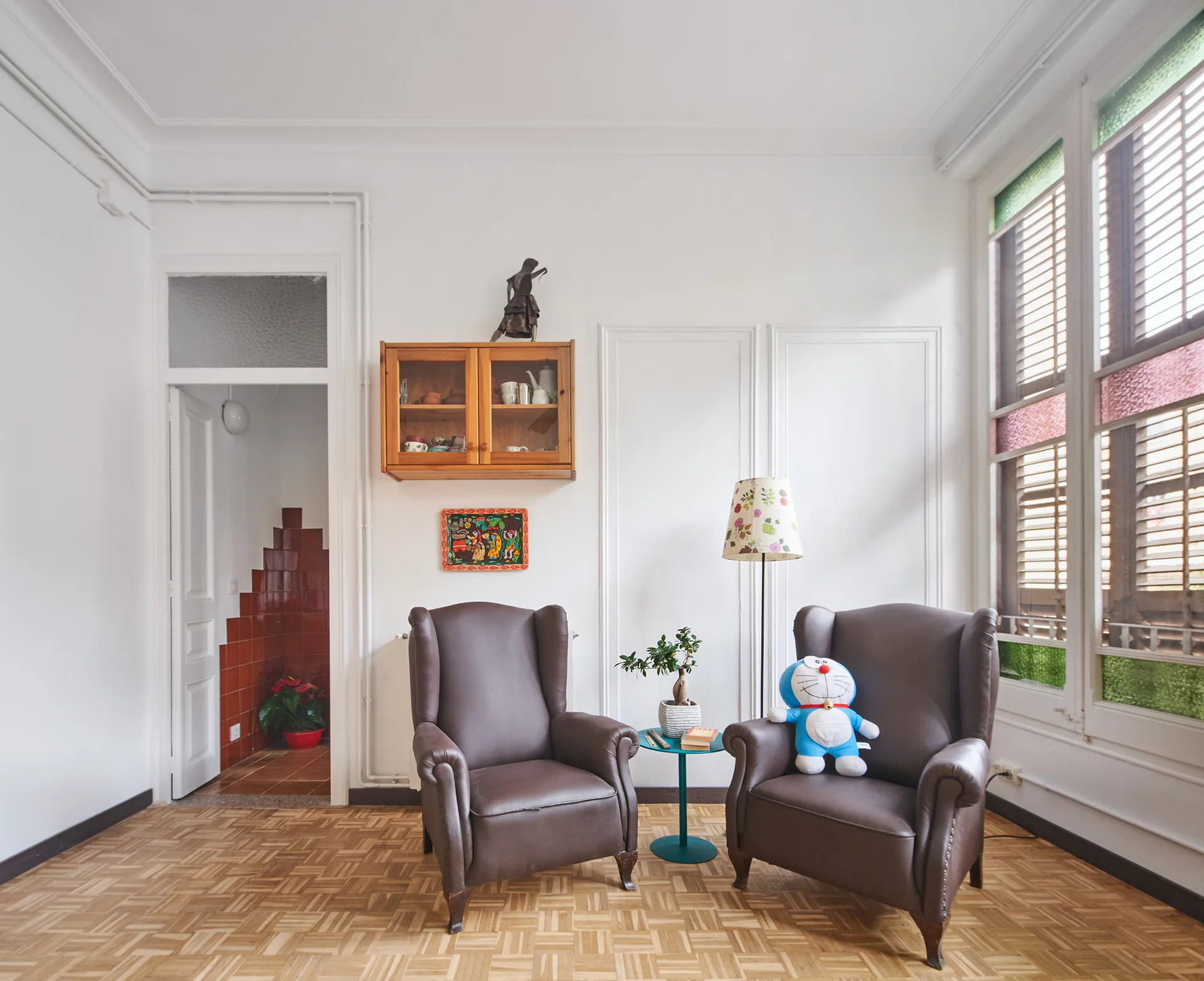 A bright living room features two brown leather wingback chairs on parquet wood flooring next to a small teal side table and a large window.