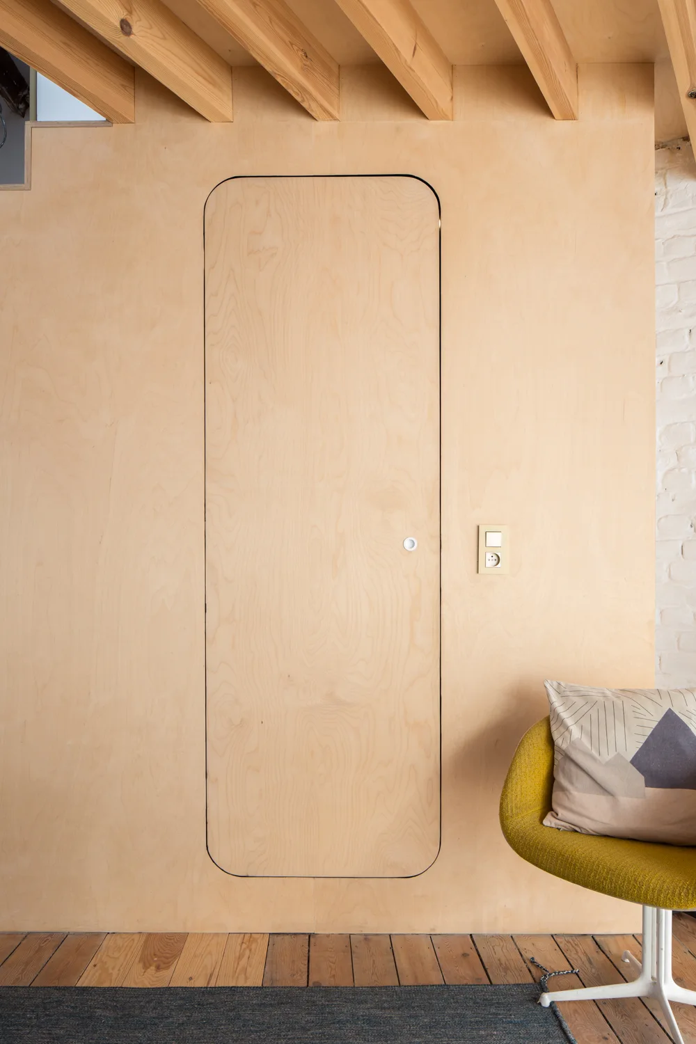 A flush-mounted plywood door with rounded corners integrated into a wood-paneled wall next to a yellow armchair and exposed ceiling beams.