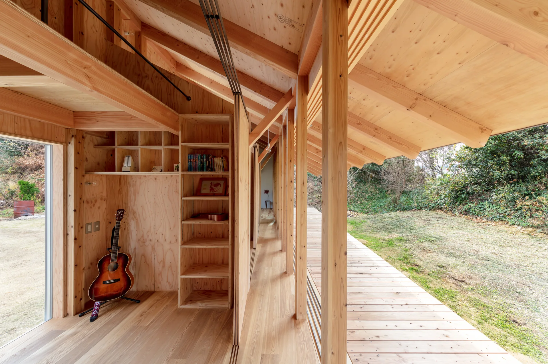 A light wood residential interior features exposed timber beams, built-in plywood shelving, a guitar, and a glass door opening to an outdoor deck.