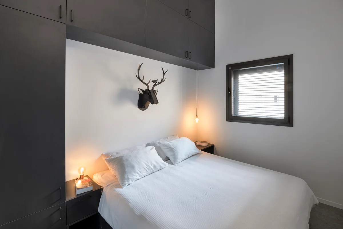 A modern bedroom featuring a bed with white linens framed by matte black built-in storage cabinets and a geometric deer head sculpture on the wall.