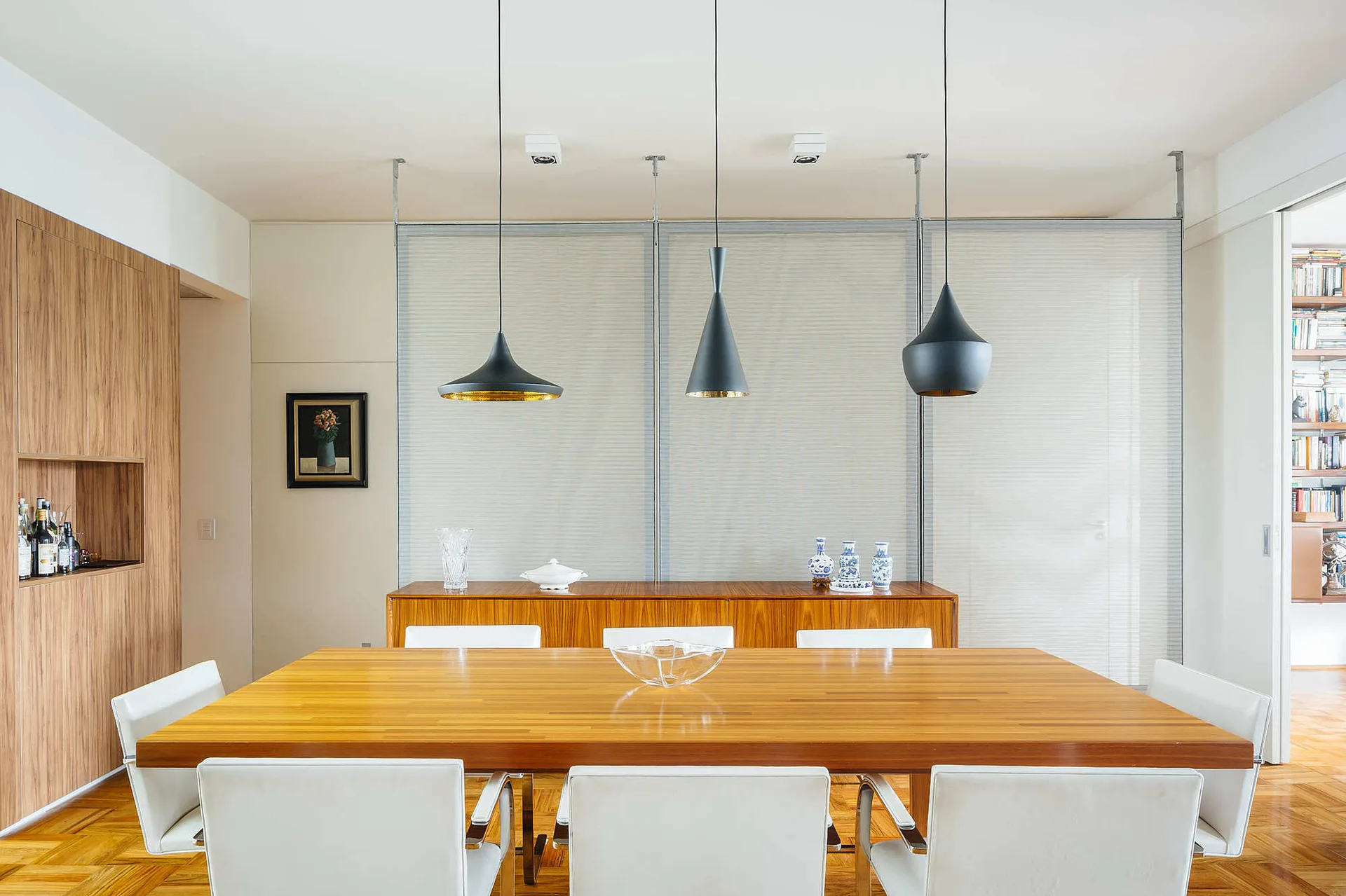 Three black pendant lights hang over a wooden dining table with white chairs in front of a warm wood sideboard and translucent sliding screens.