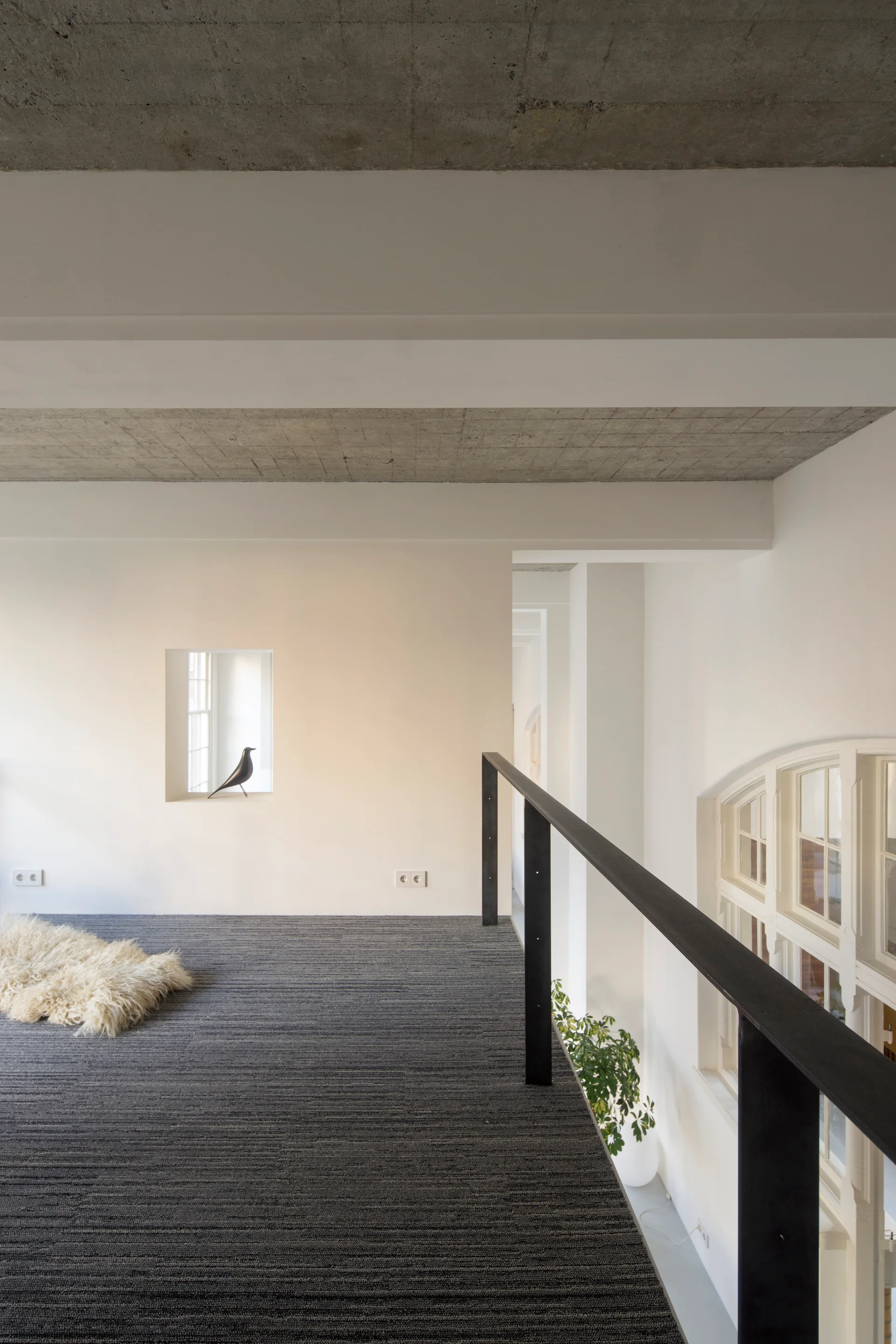 A minimalist loft landing features a dark grey carpeted floor, a black metal railing, white walls, and an exposed concrete ceiling.