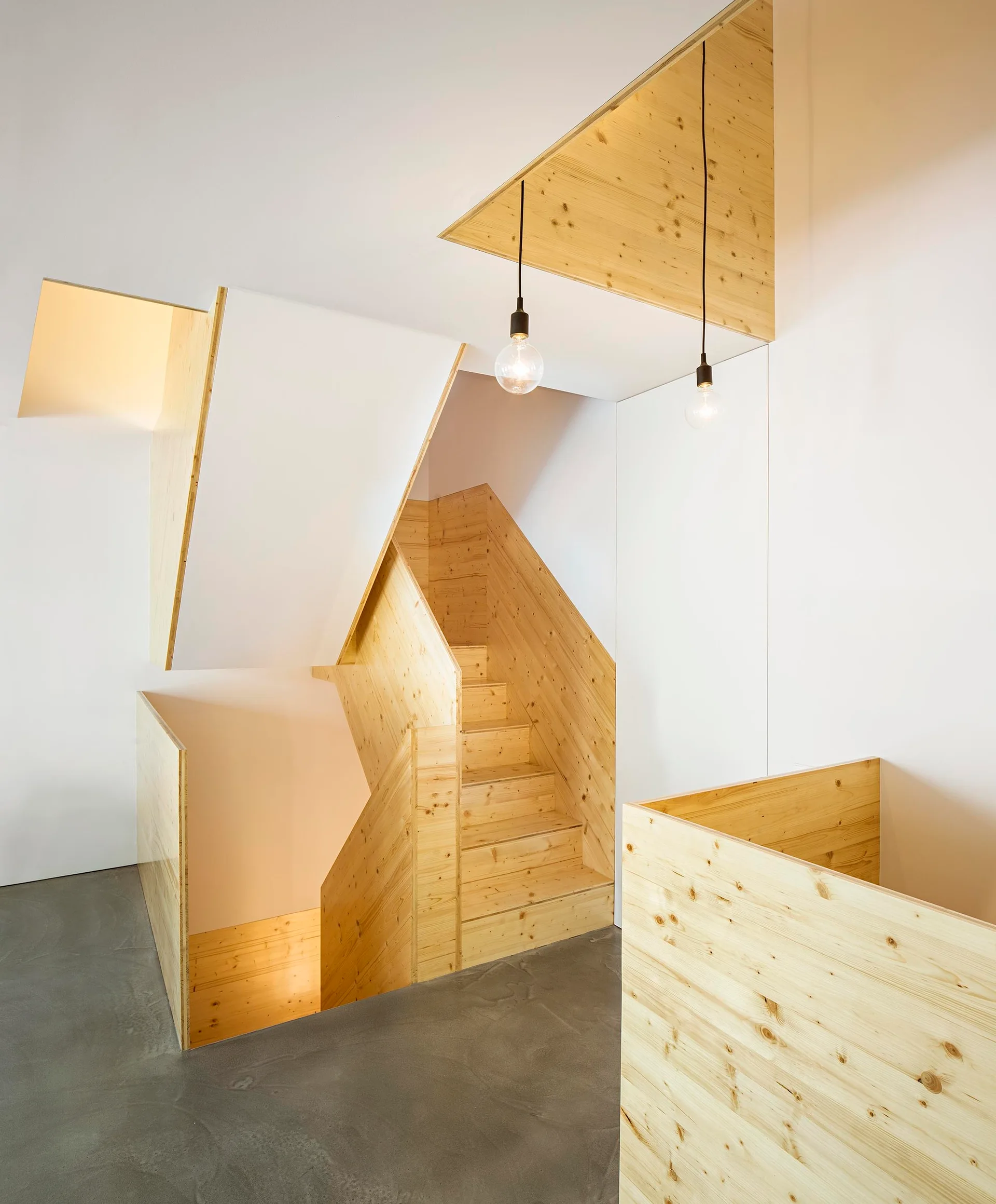 A minimalist staircase crafted from light wood is set against smooth white walls and a gray concrete floor with pendant light bulbs.