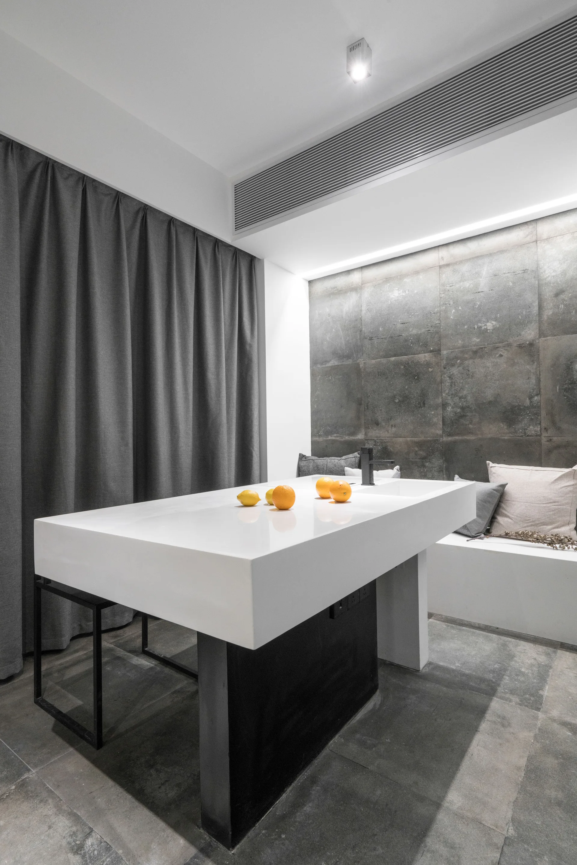 A white minimalist utility island with an integrated sink and black faucet stands in front of a dark tiled wall and a built-in bench.
