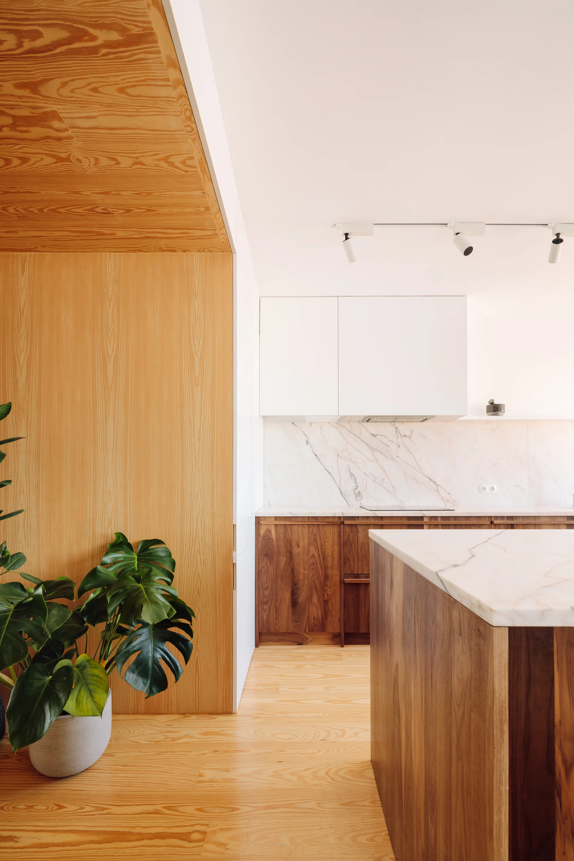 A minimalist kitchen features a marble-topped island, dark wood cabinets, a marble slab backsplash, white upper cabinets, and light wood wall cladding.