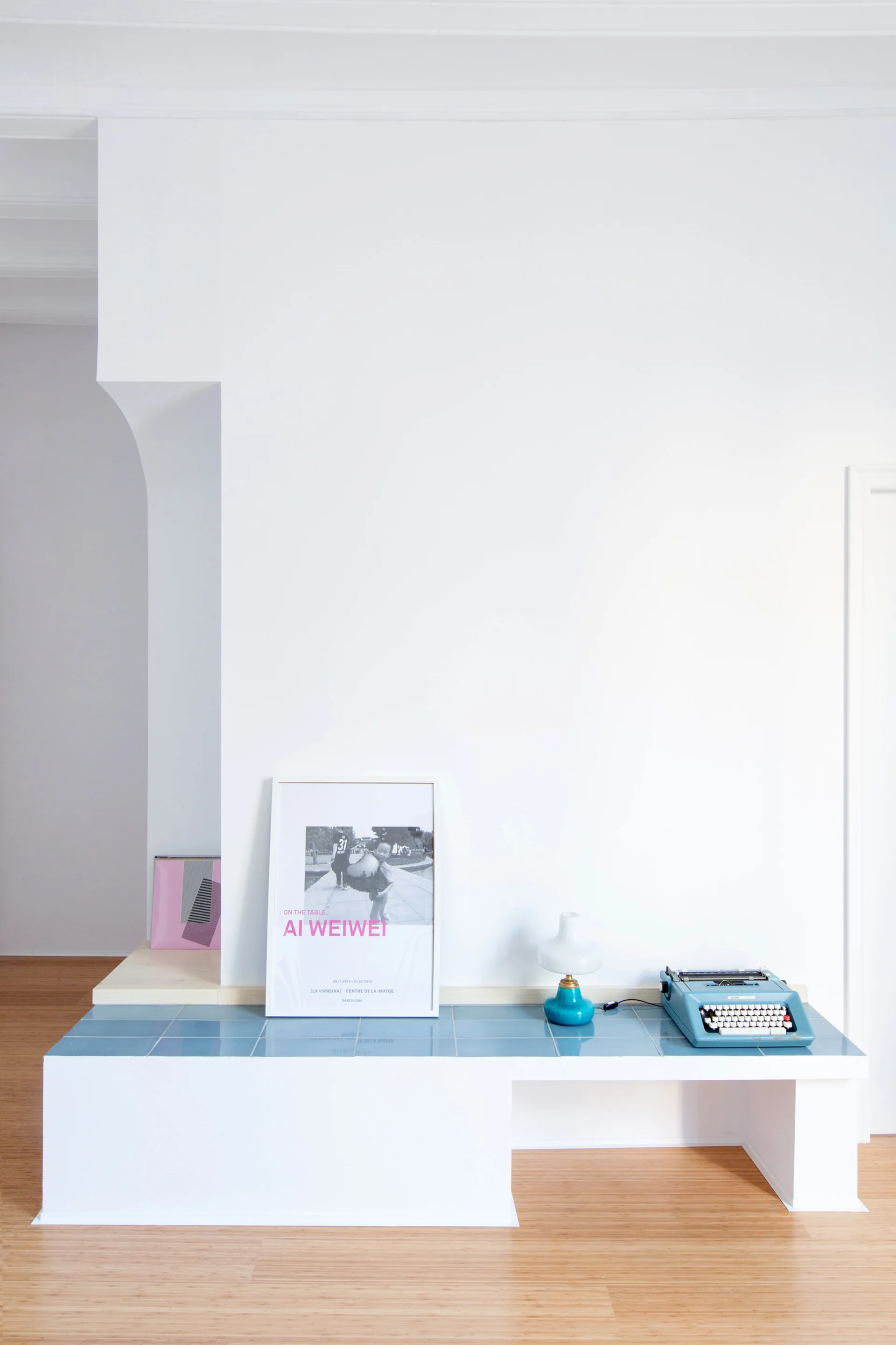 A minimalist built-in white bench with a blue tiled top features a framed poster, a small lamp, and a vintage typewriter against a white wall.