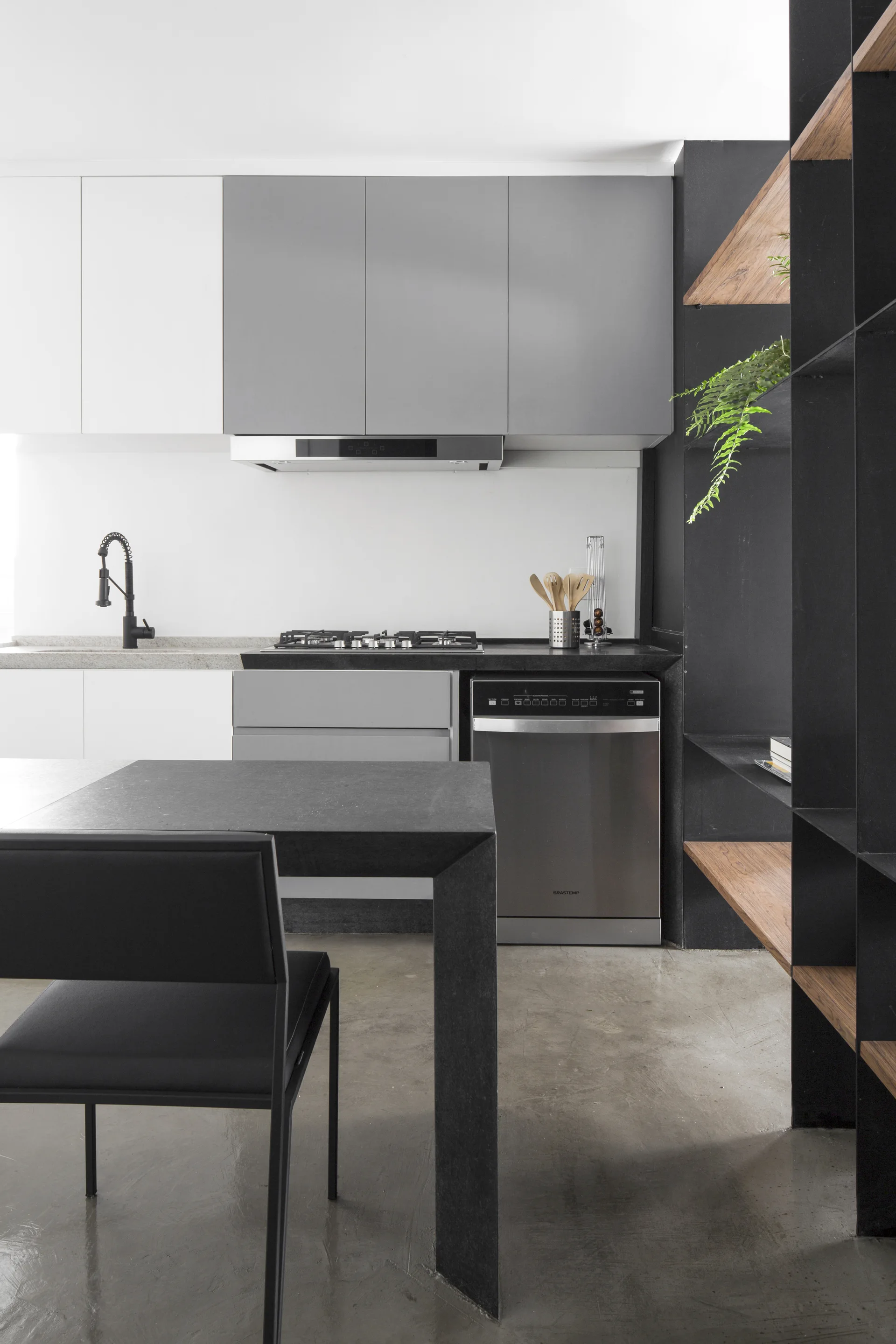 A modern kitchen featuring grey and white cabinetry, a black dining table, and a black metal shelving unit with wood shelves on a concrete floor.