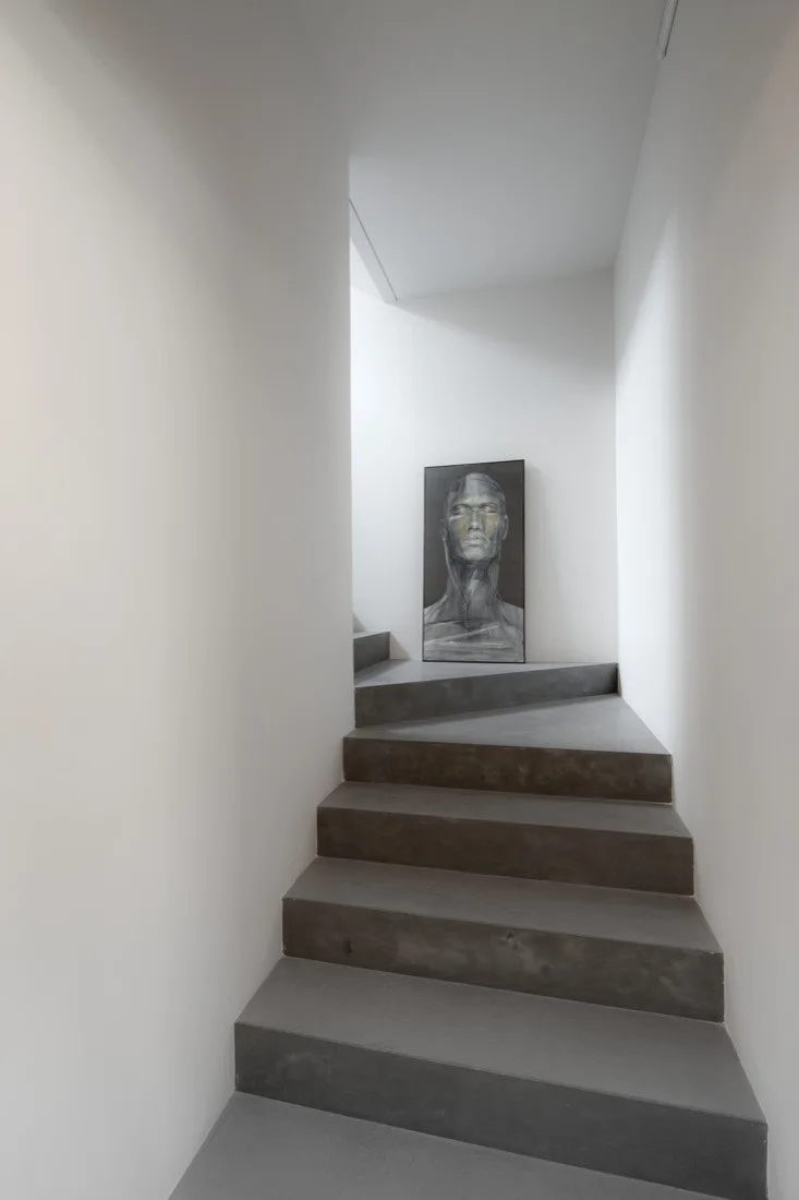 Dark concrete stairs wind upward between smooth white walls toward a landing decorated with a vertical black-and-white portrait painting.