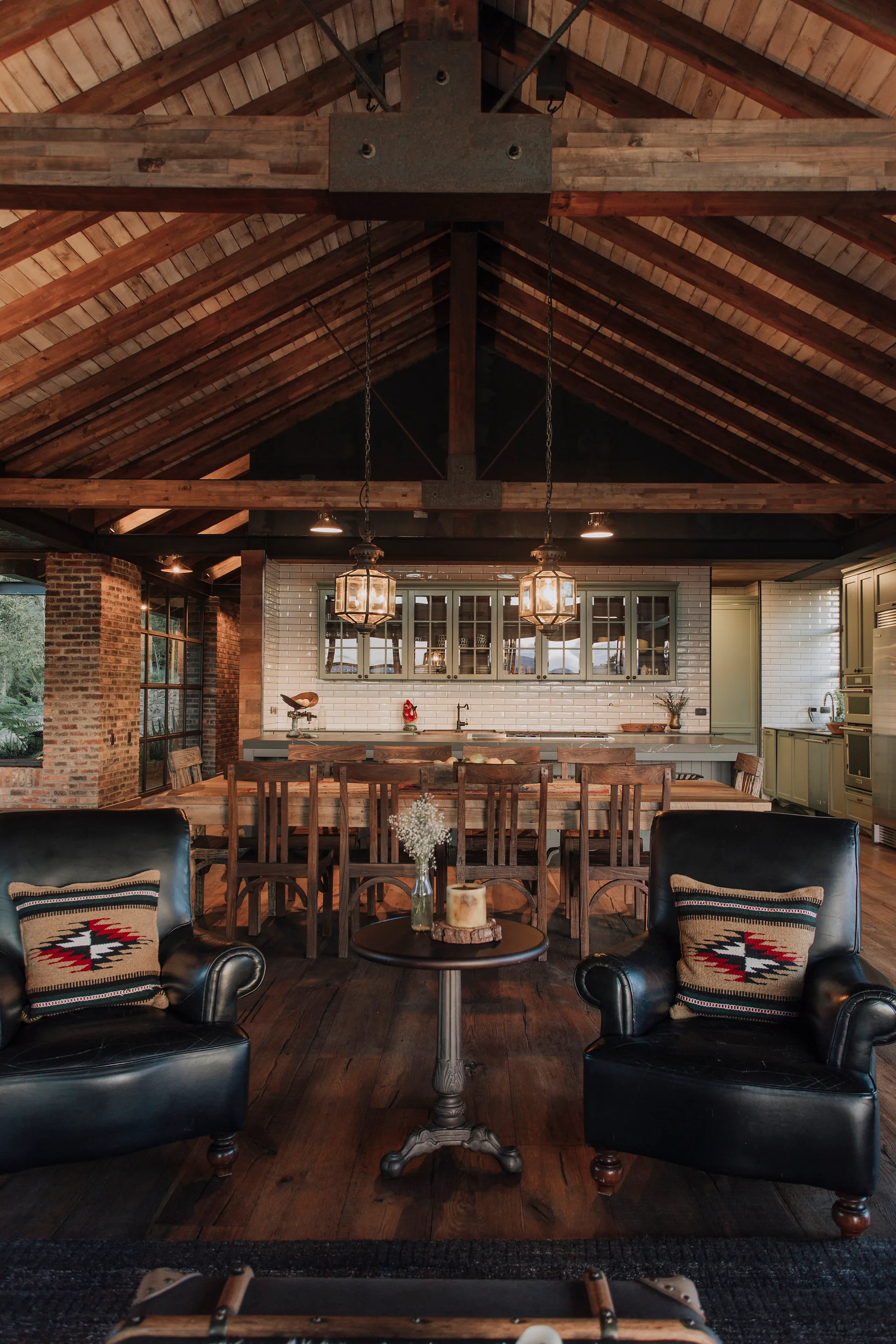 A rustic dining room featuring a high vaulted wood ceiling with exposed beams, a large wooden table, and two black leather armchairs.