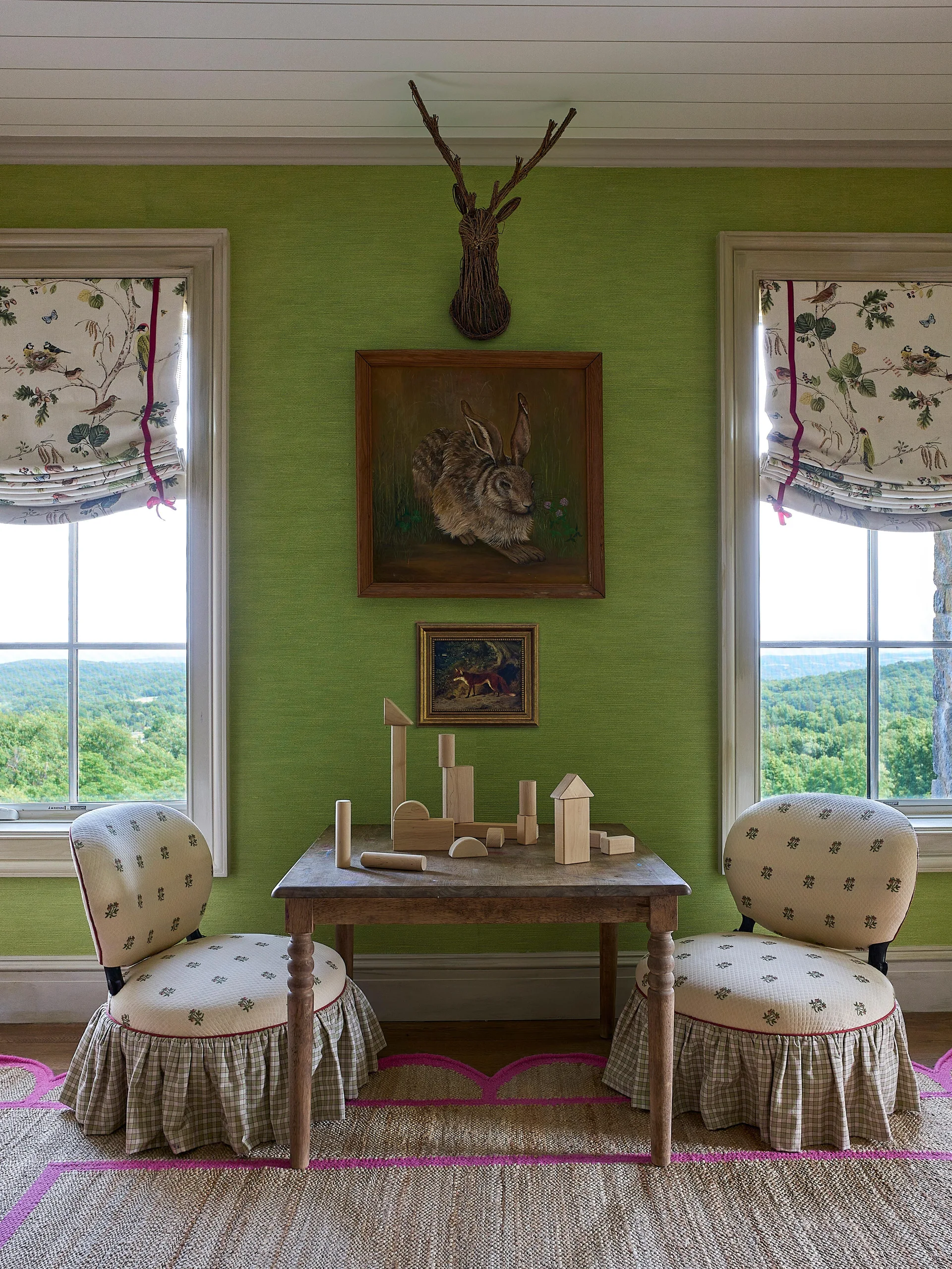 A children's wooden play table with blocks sits between two patterned skirted chairs against a bright green wallpapered wall with windows.