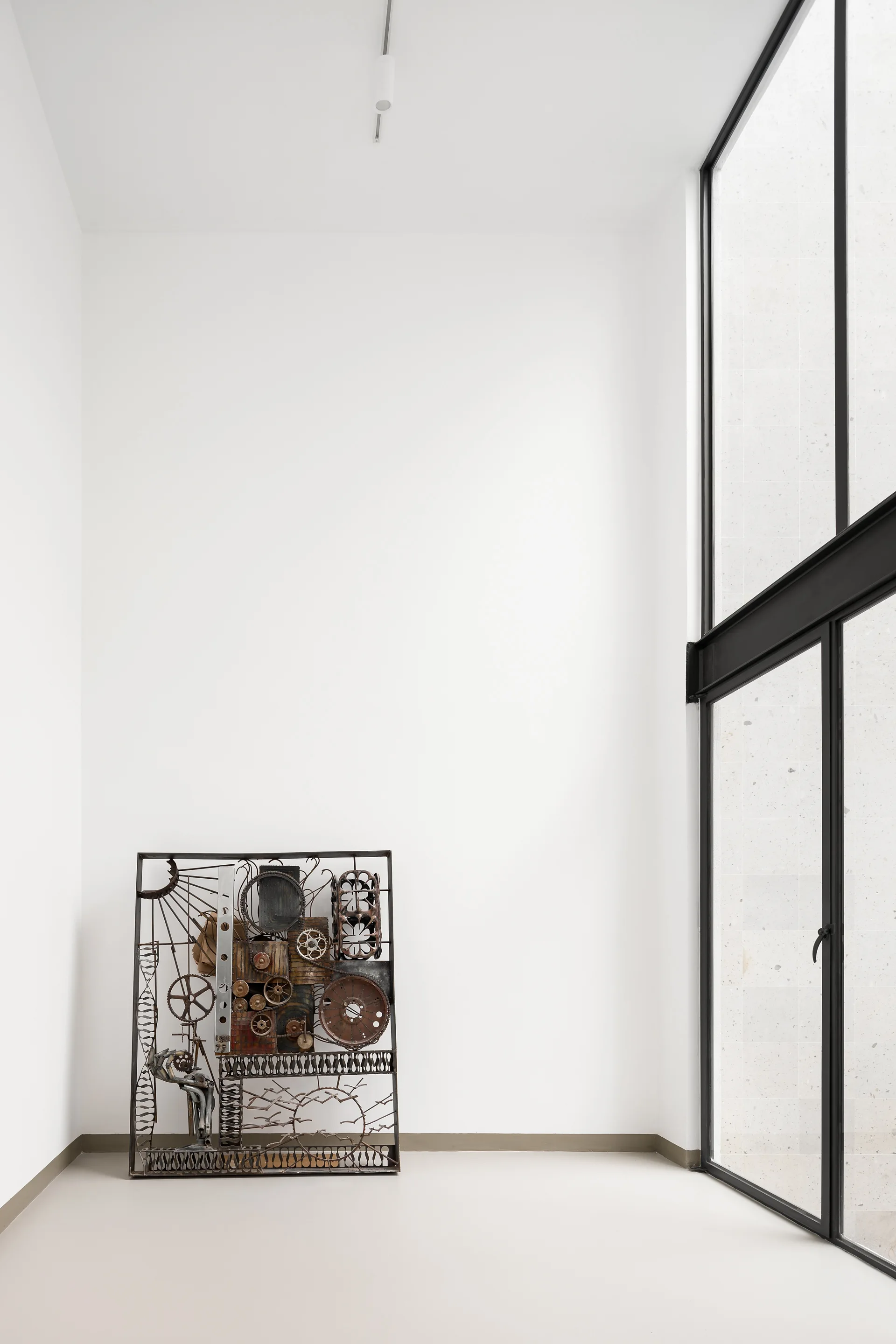A tall minimalist room with white walls features a rectangular industrial metal sculpture on the floor beside a large black-framed glass door.