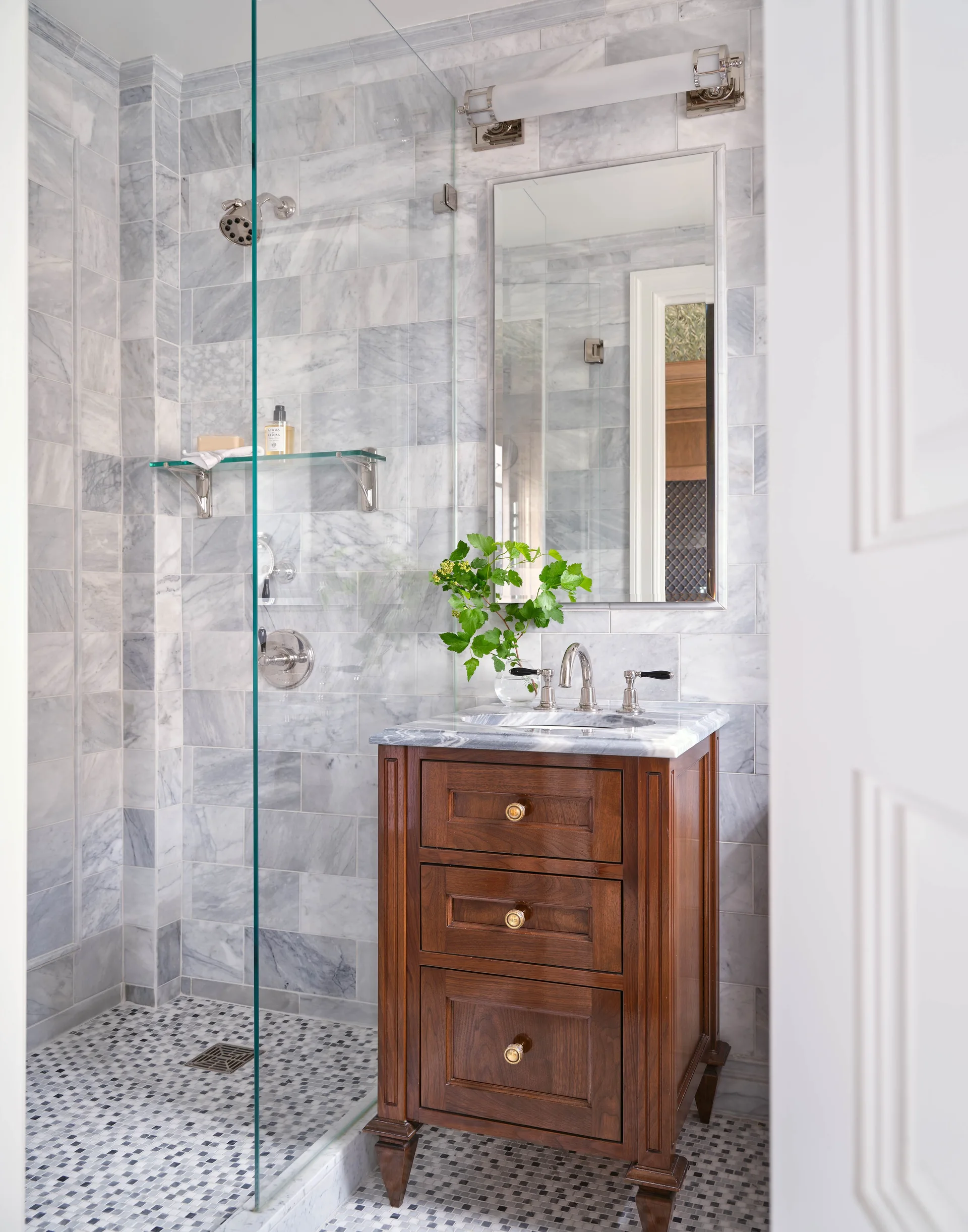 A bathroom features grey marble wall tiles, a glass-enclosed walk-in shower, a mosaic tile floor, and a dark wood vanity with brass hardware.