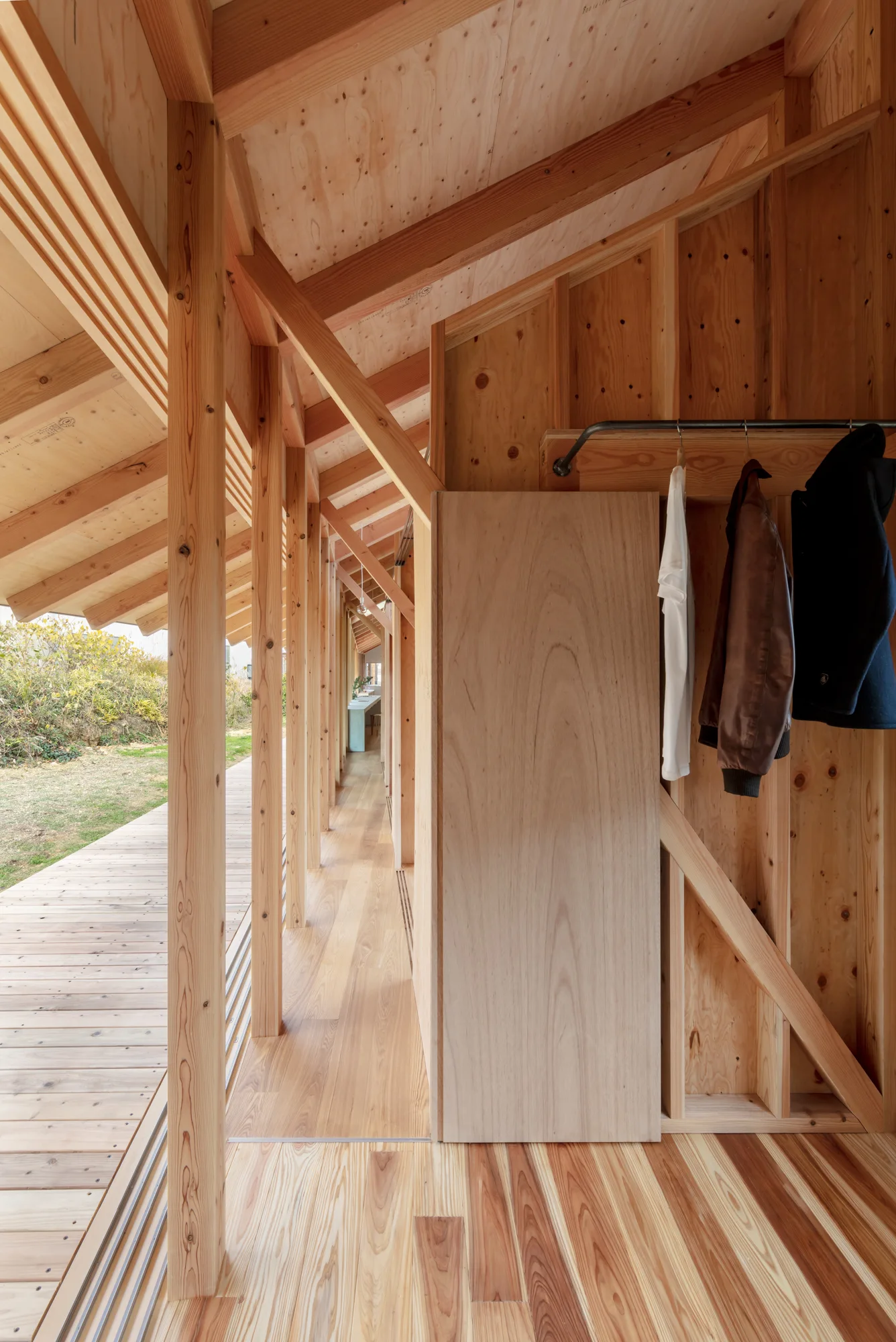 A light-filled wooden hallway featuring an open closet with hanging clothes, plywood walls, and an exposed timber frame.