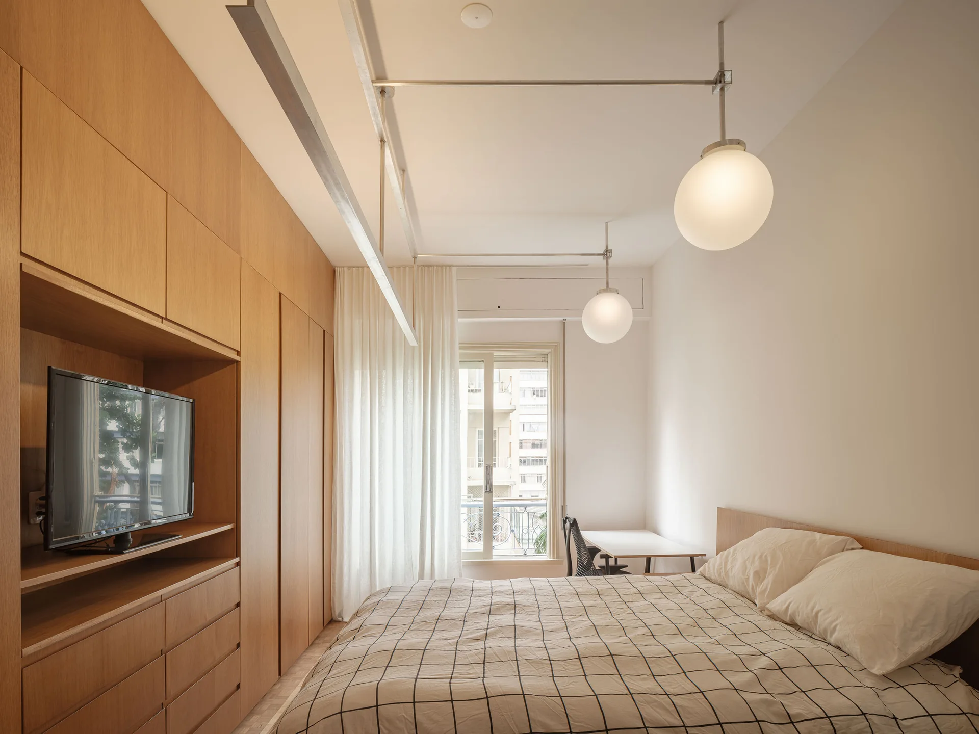 A minimalist bedroom features a floor-to-ceiling wood wardrobe with an integrated TV, a bed with grid-patterned bedding, and sphere pendant lights.