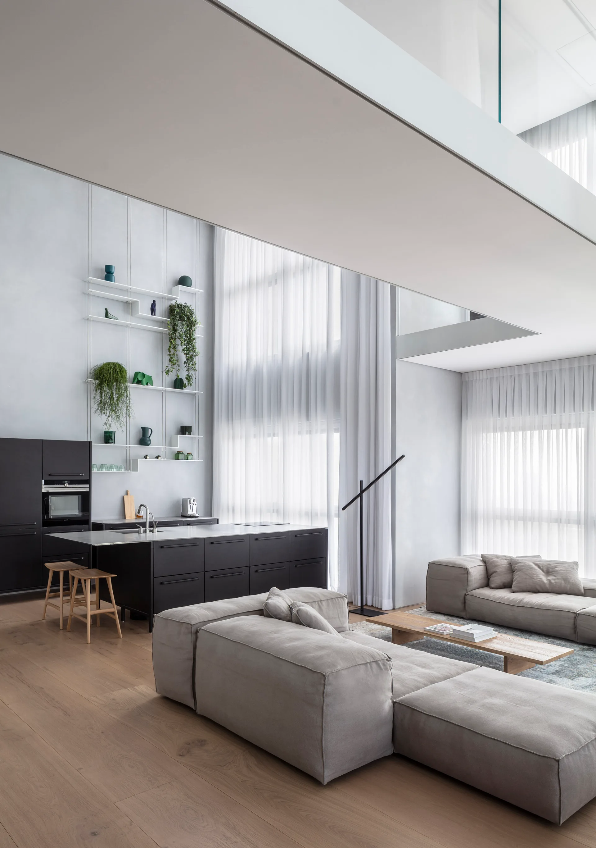 An open-plan living space features a large grey modular sofa, a black kitchen island, and tall windows with sheer white curtains.