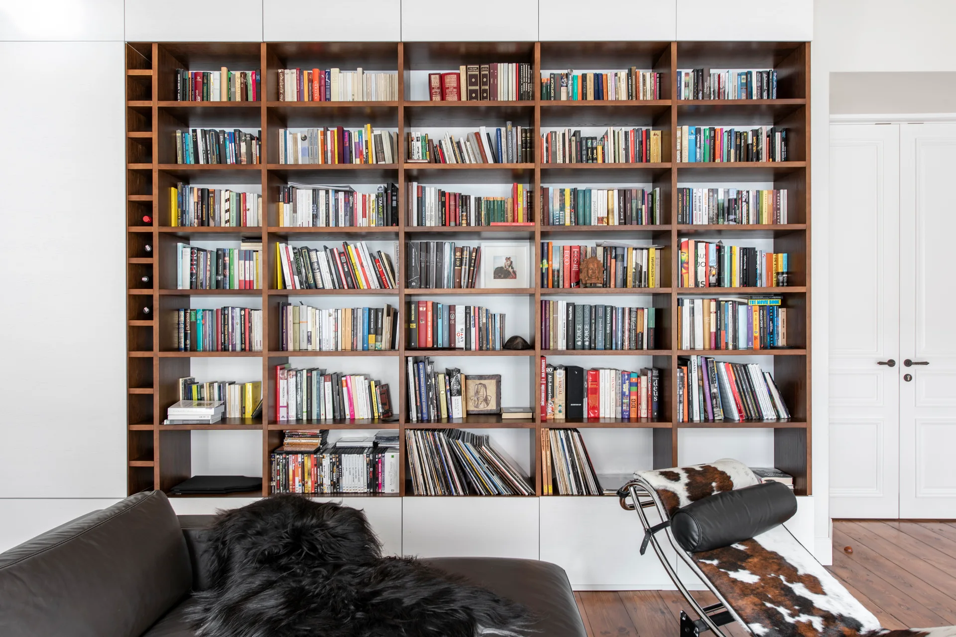 A dark wood grid bookcase integrated into a white wall is filled with books and vinyl records, near a black leather sofa and a cowhide chaise.