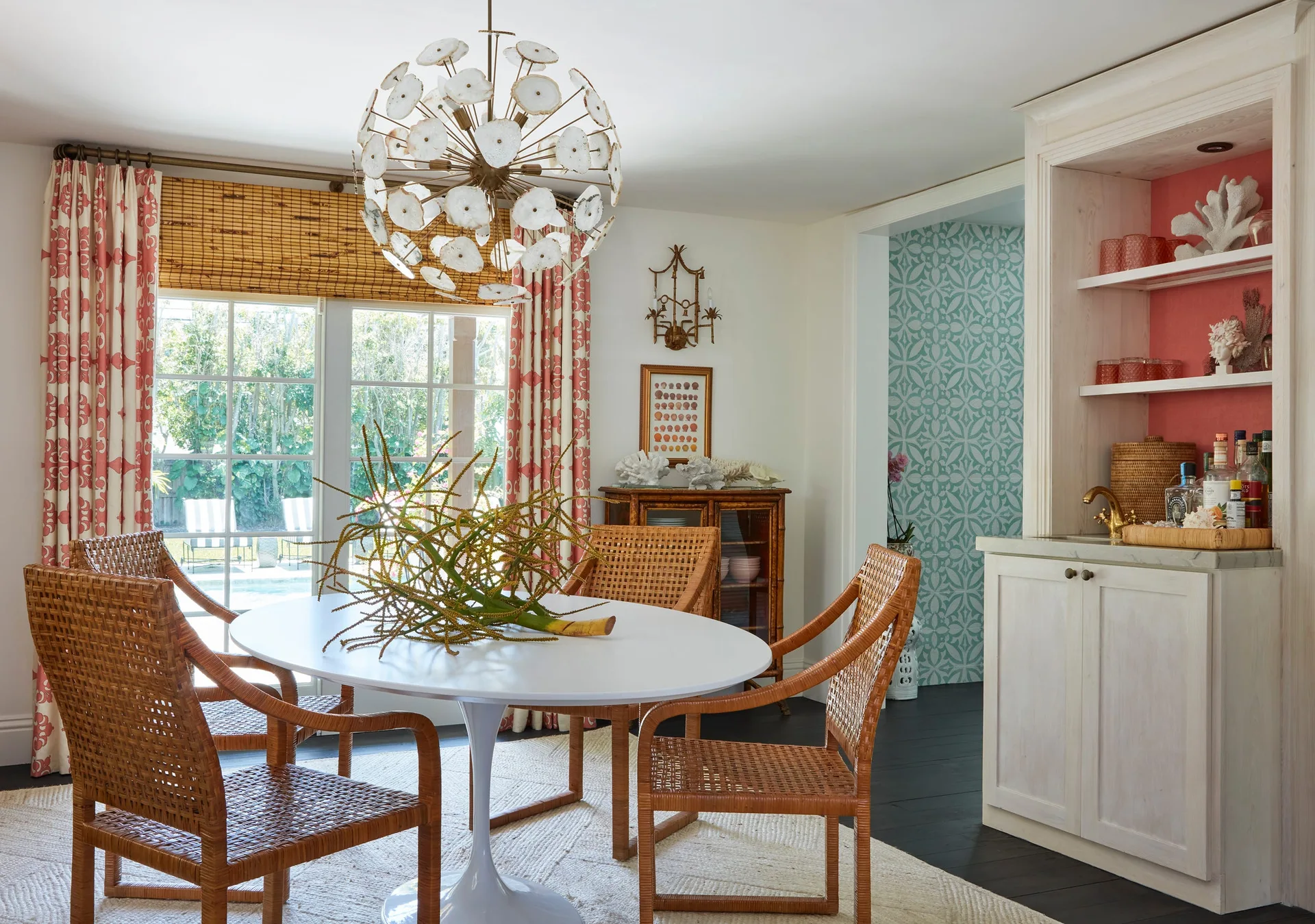 A coastal dining room featuring a white round tulip table, woven wicker chairs, a capiz shell globe chandelier, and a built-in wet bar.