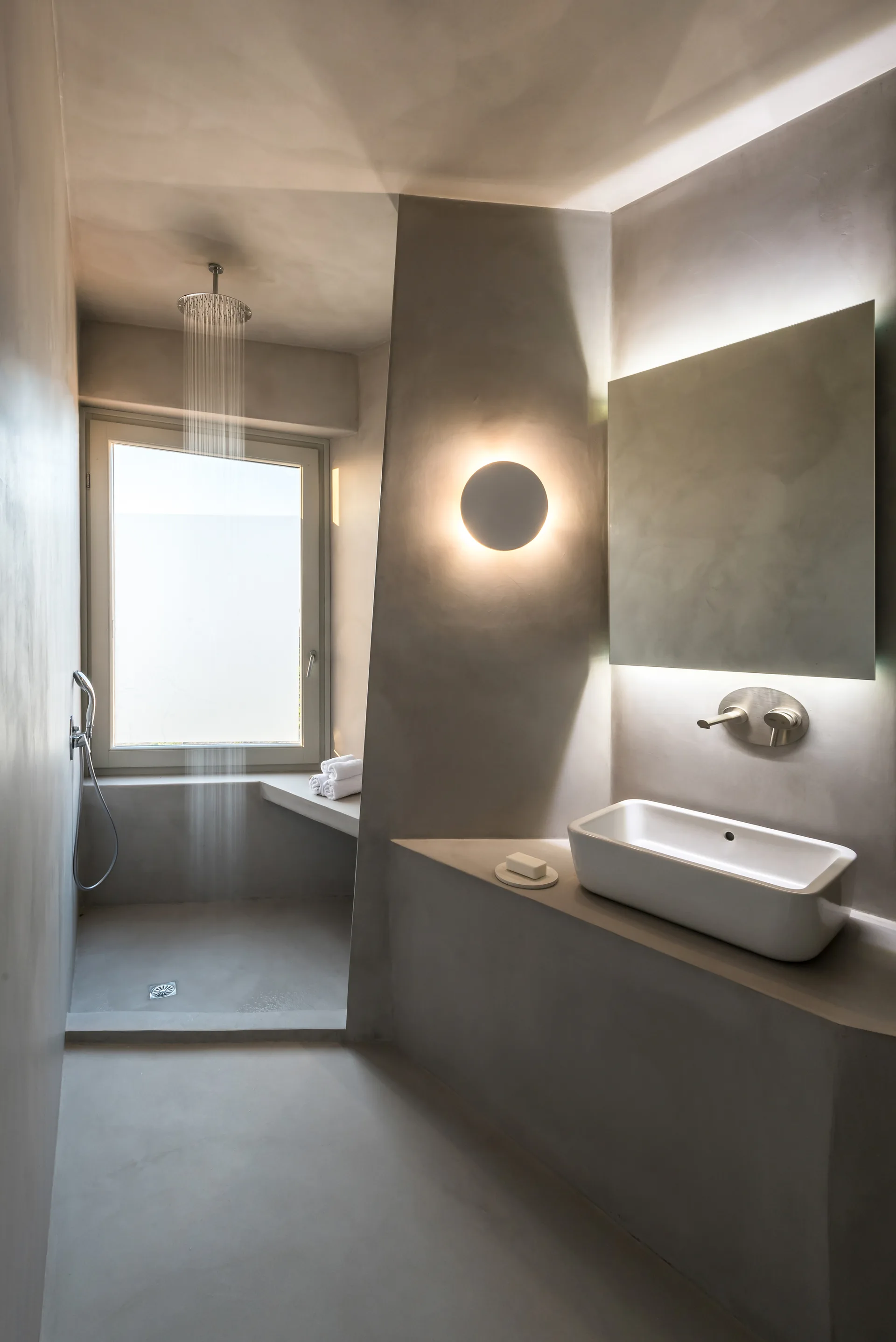 A minimalist gray plaster bathroom features a built-in vanity with a white vessel sink, a backlit mirror, and an open walk-in shower.