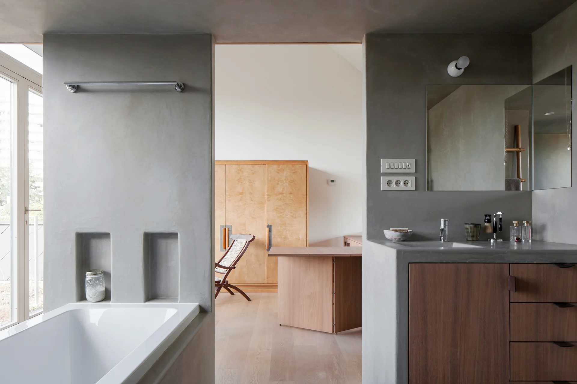 A modern grey plaster bathroom features a built-in tub with niches and a vanity, looking into a bedroom with wooden furniture.
