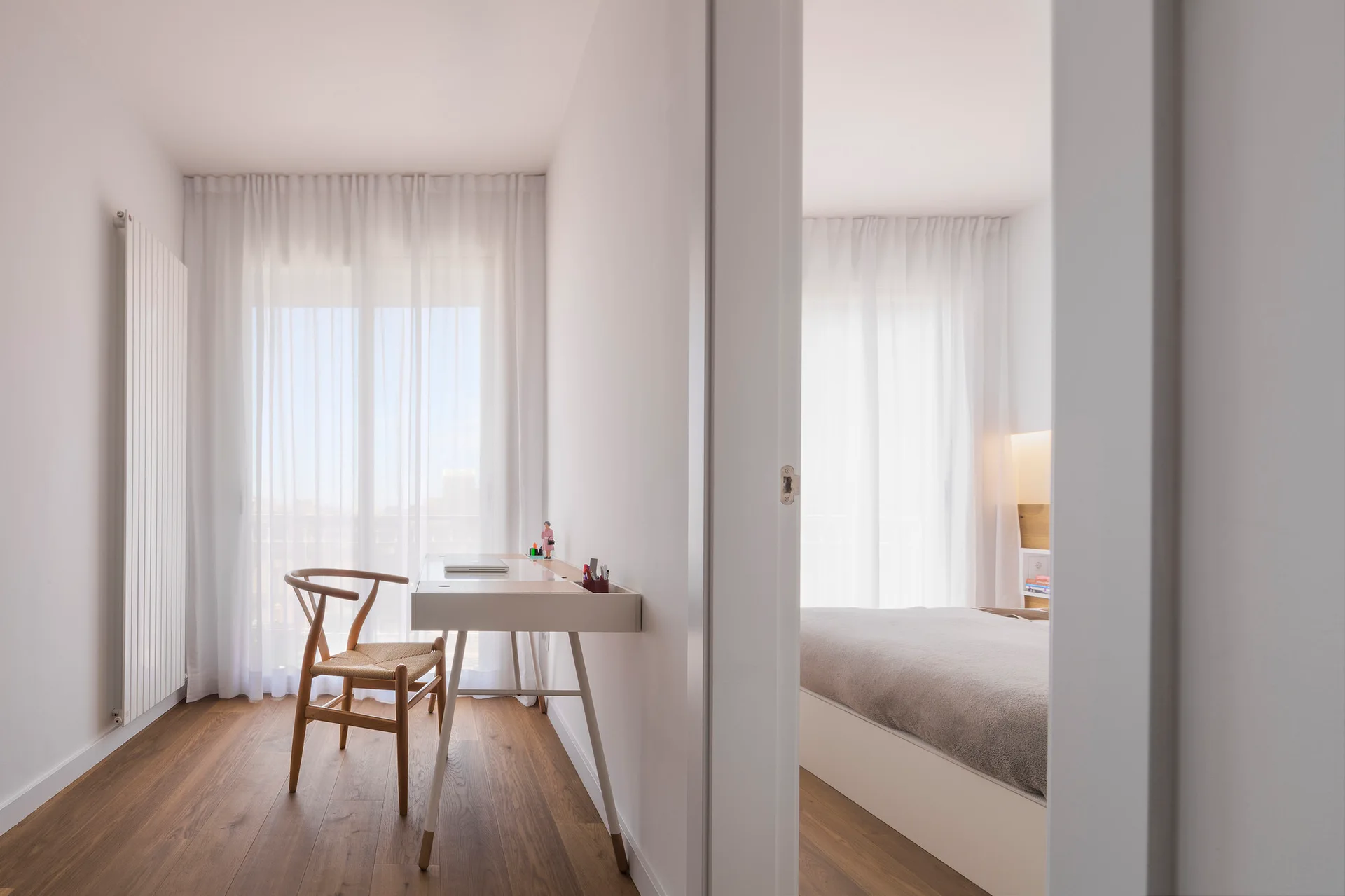 A minimalist home office featuring a white desk and a wooden wishbone chair next to an open doorway leading to a bedroom.