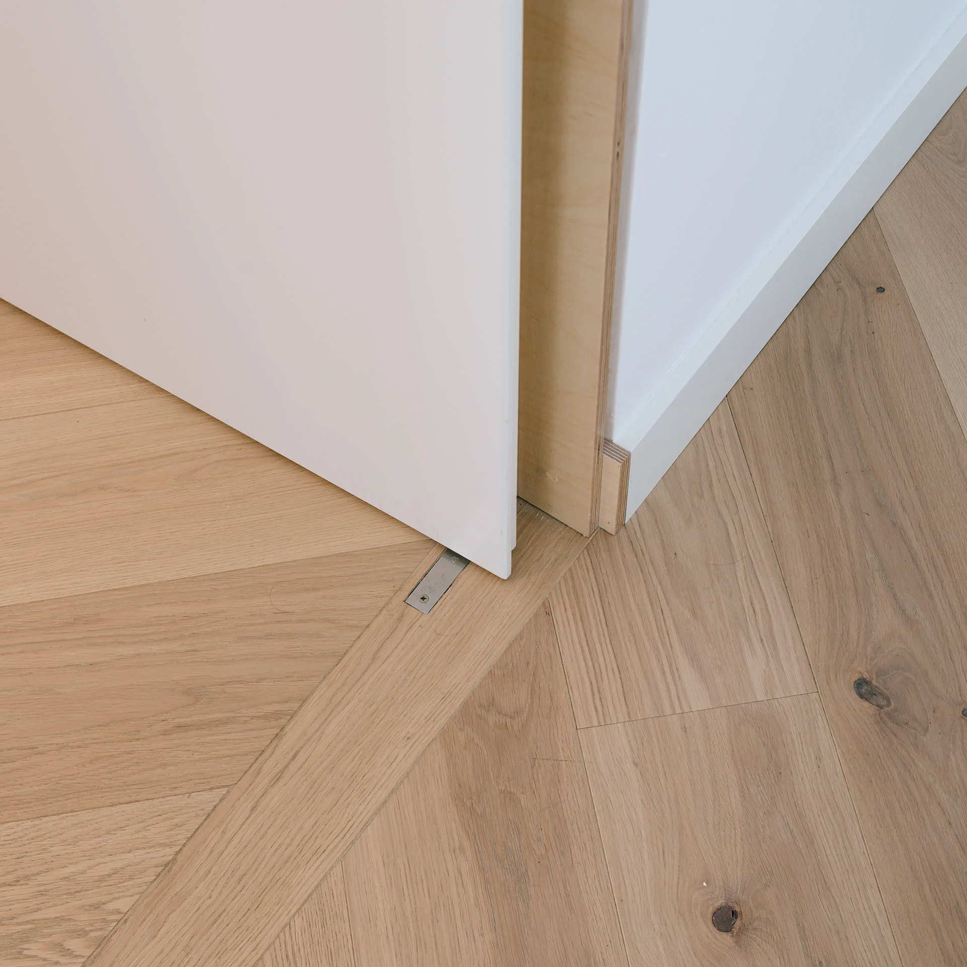 Close-up of a white door and an exposed plywood door jamb meeting a light oak herringbone pattern wood floor.