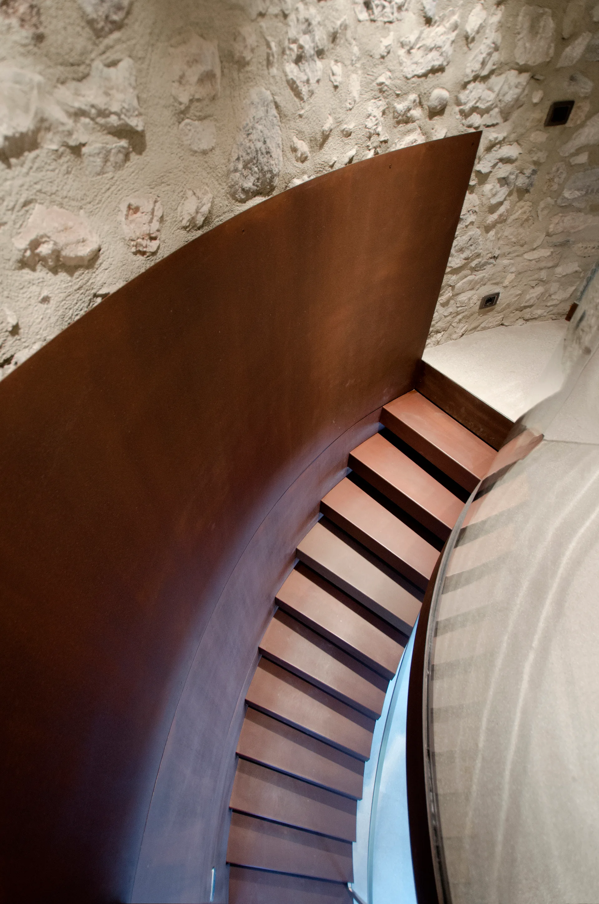 An overhead view of a curved metal staircase with rust-colored treads set against a textured, light beige exposed stone wall.