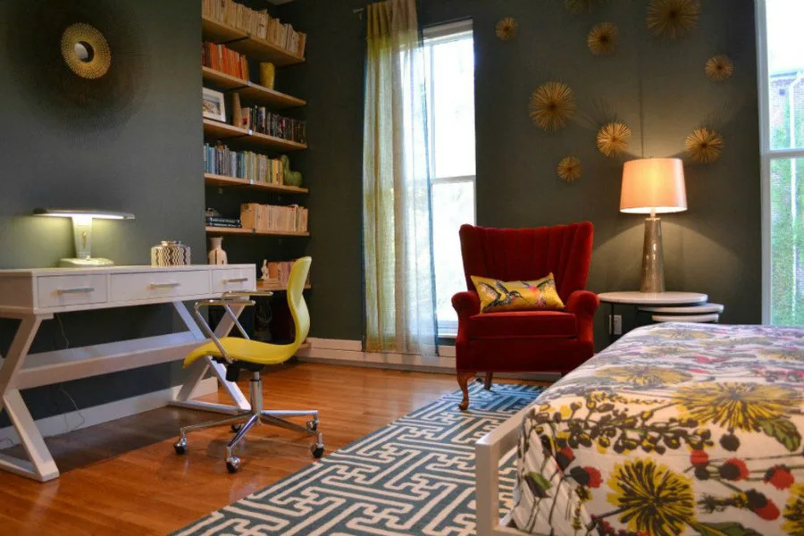 An eclectic bedroom features deep green walls, a white writing desk with a yellow chair, a red armchair, and built-in wooden bookshelves.