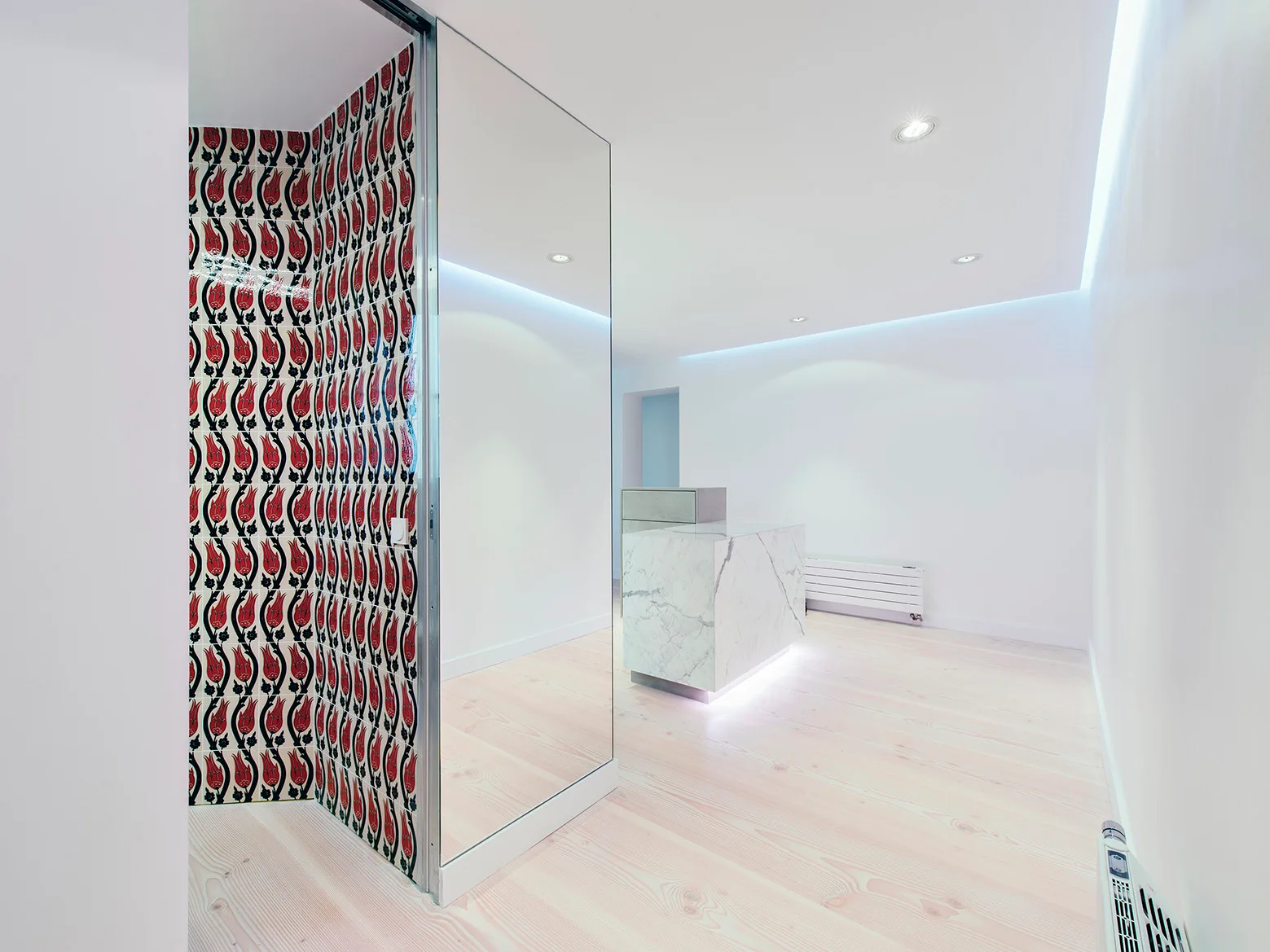 A minimalist lobby features a marble reception counter, light wood floors, a tall mirror, and an accent wall tiled in a red and black tulip pattern.