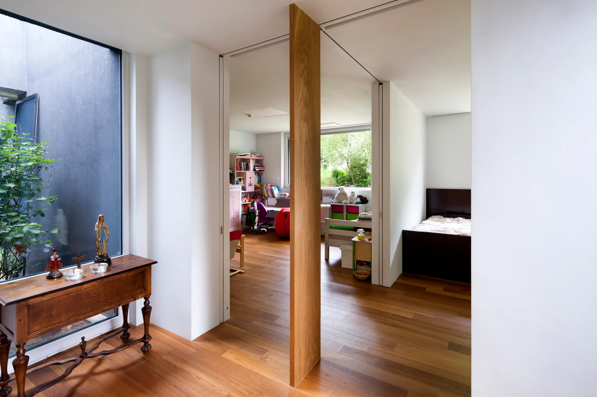 A bright residential interior features hardwood floors, white walls, a slim vertical wood partition, and doorways leading to a bedroom and children's room.