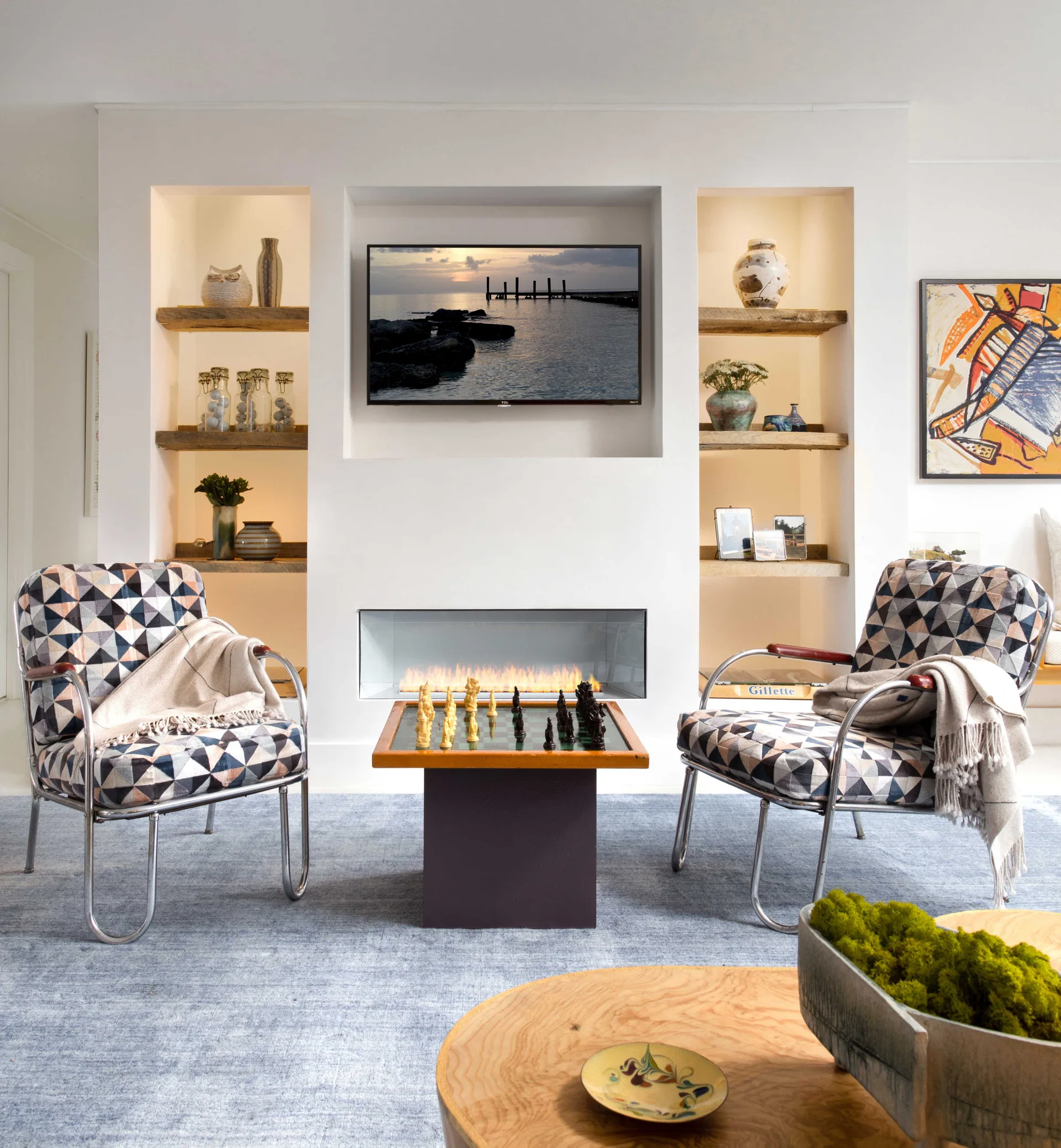 Two geometric pattern armchairs flank a small chess table in front of a modern linear fireplace with built-in wooden shelving niches.