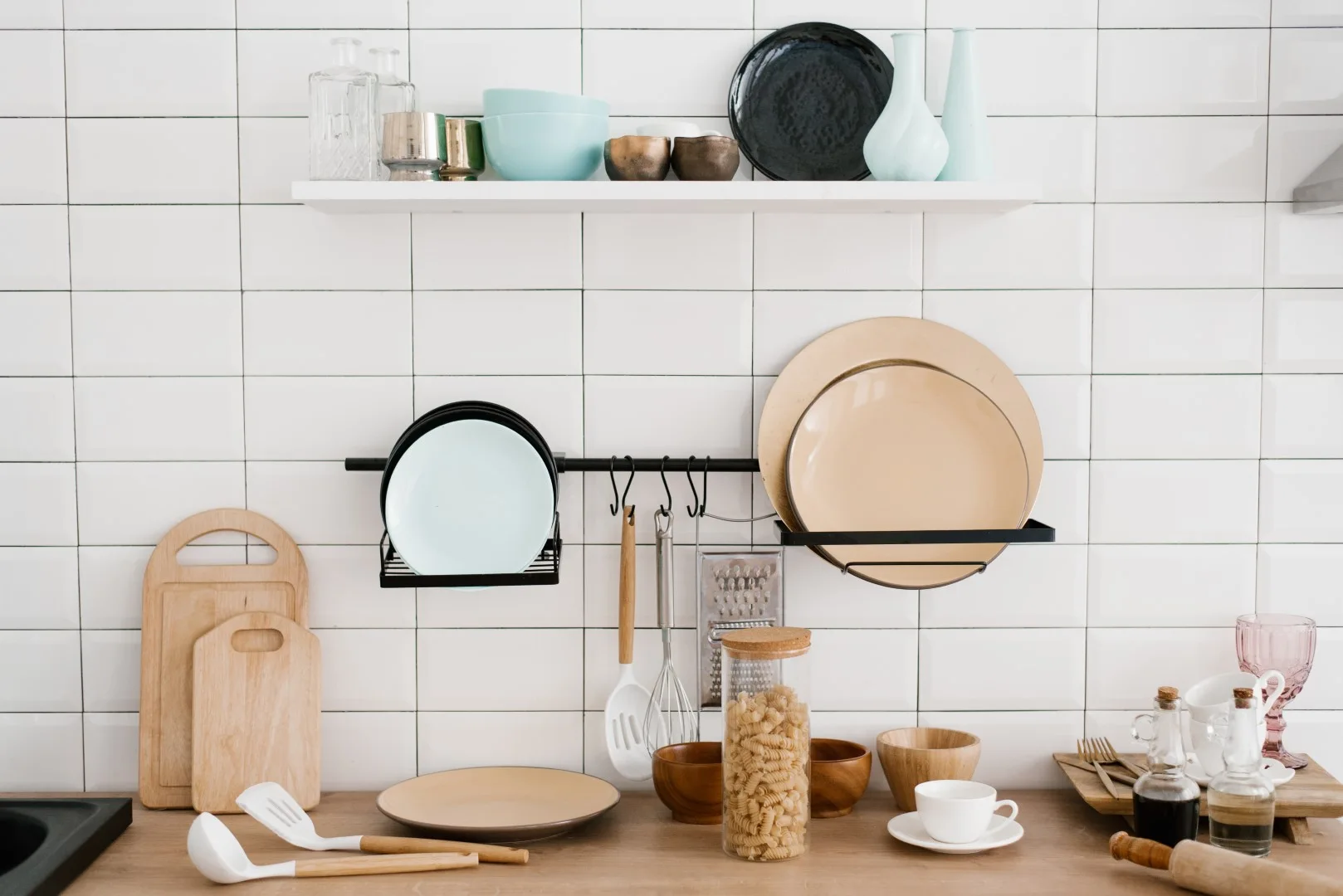 A kitchen counter with a white subway tile backsplash, a white floating shelf, and a black metal wall rail holding plates and utensils.