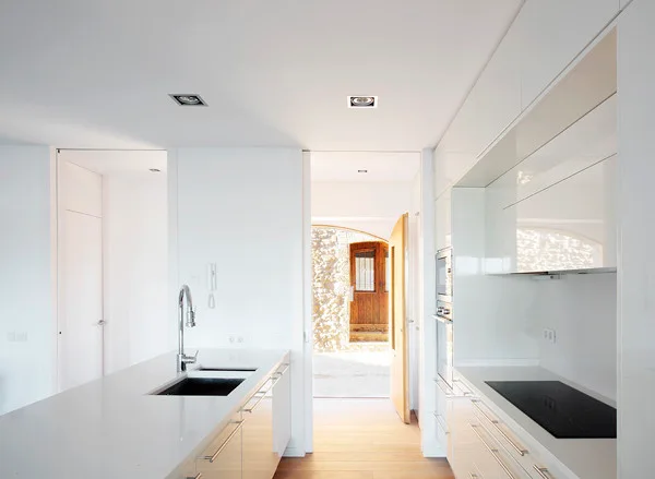 A minimalist white kitchen featuring glossy cabinets, a central island with a sink, and an open doorway leading to a stone wall exterior.