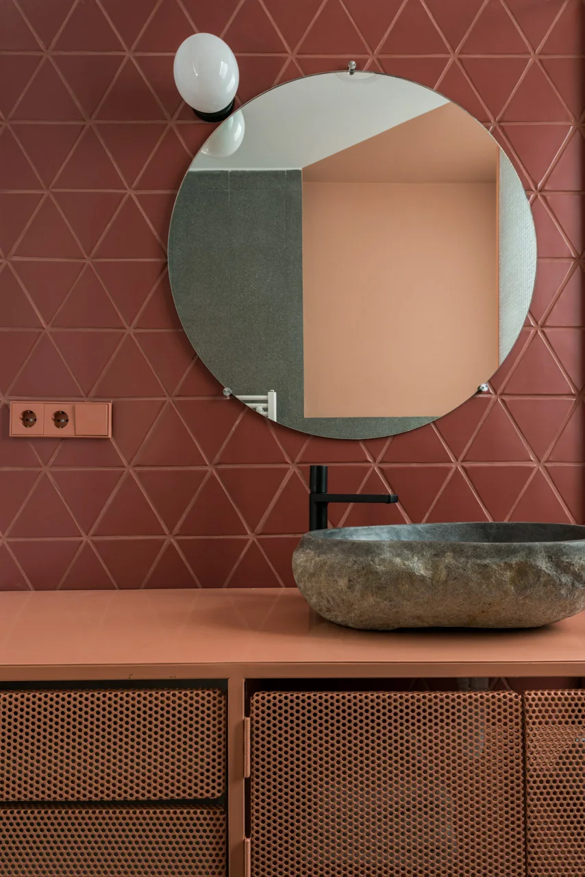 A terracotta bathroom wall with triangular tiles, a round mirror, and a rough stone vessel sink sitting on a matching perforated metal vanity.