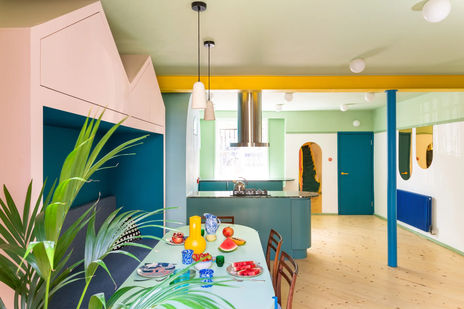 A colorful kitchen and dining area features a pink house-shaped booth, a teal island, and a light green dining table set with fruit.