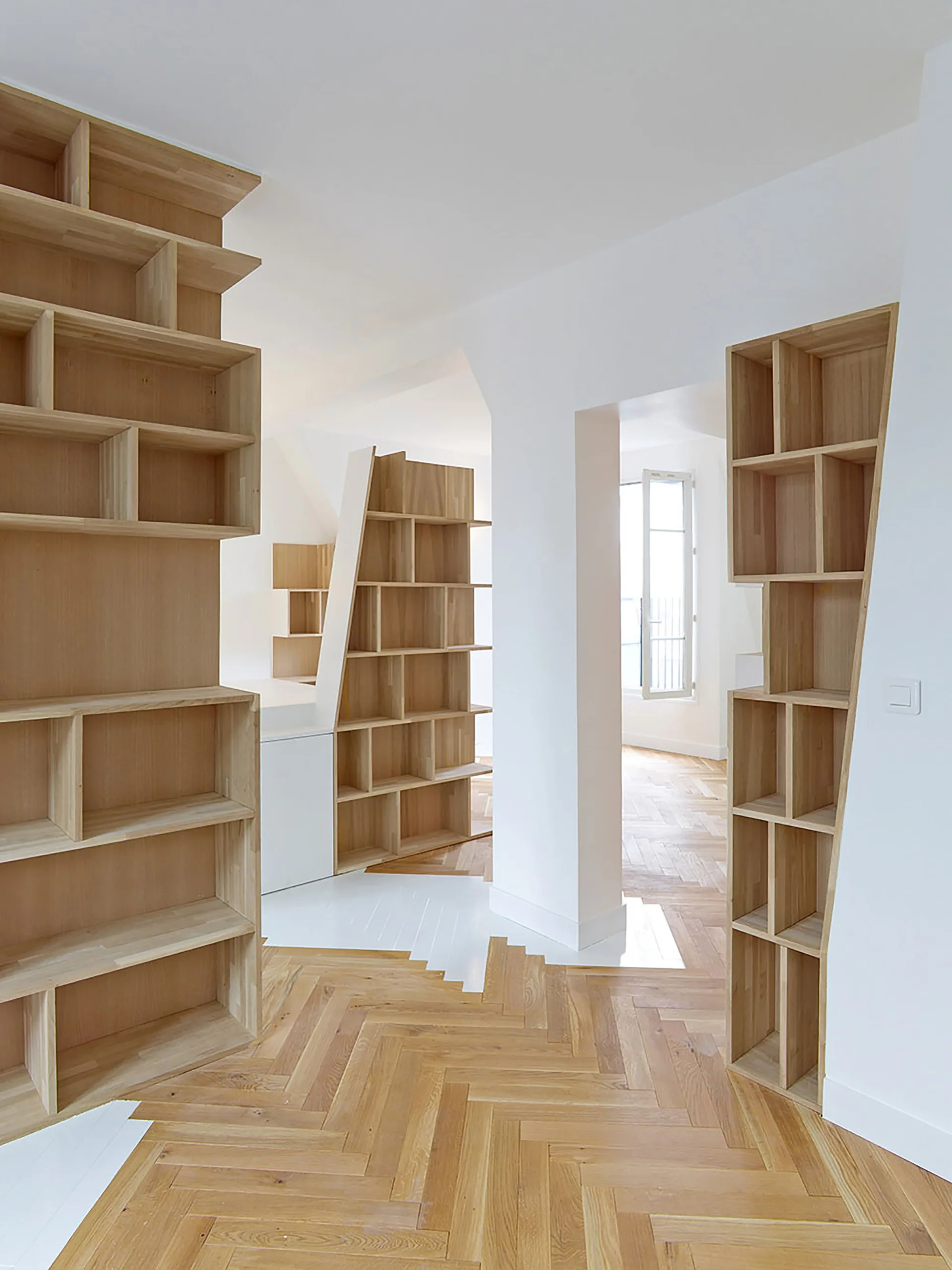 Multiple tall, custom-angled light wood bookcases line the walls of an open interior space featuring white walls and oak herringbone parquet flooring.