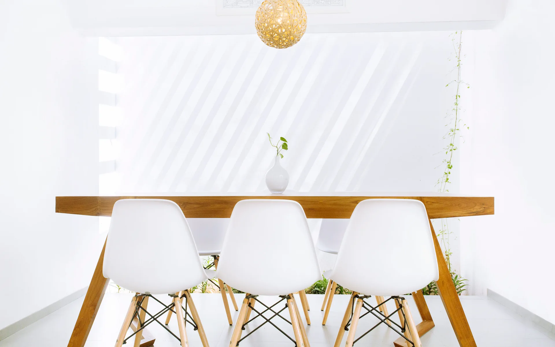 Three white molded dining chairs sit at a natural wood table beneath a woven sphere pendant light in a bright room.