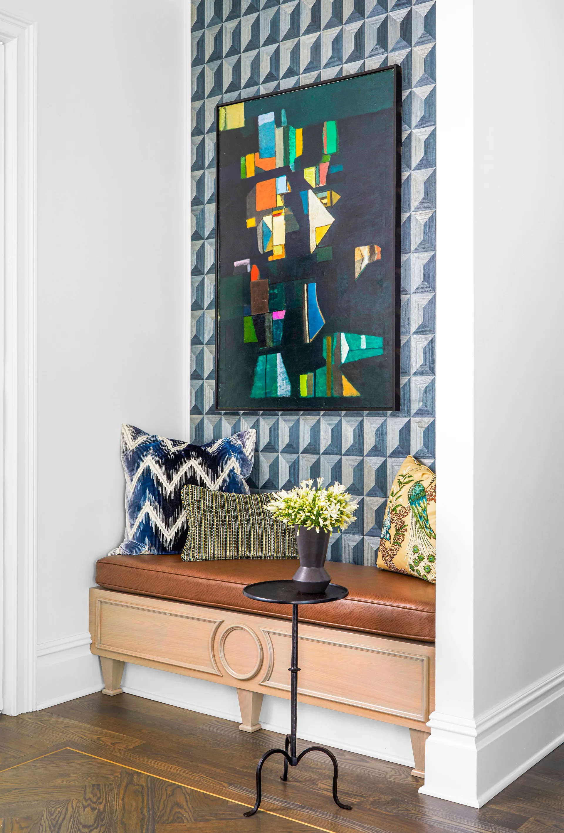 A wooden built-in bench with a brown leather cushion is recessed in an alcove lined with blue geometric wallpaper and topped with abstract artwork.