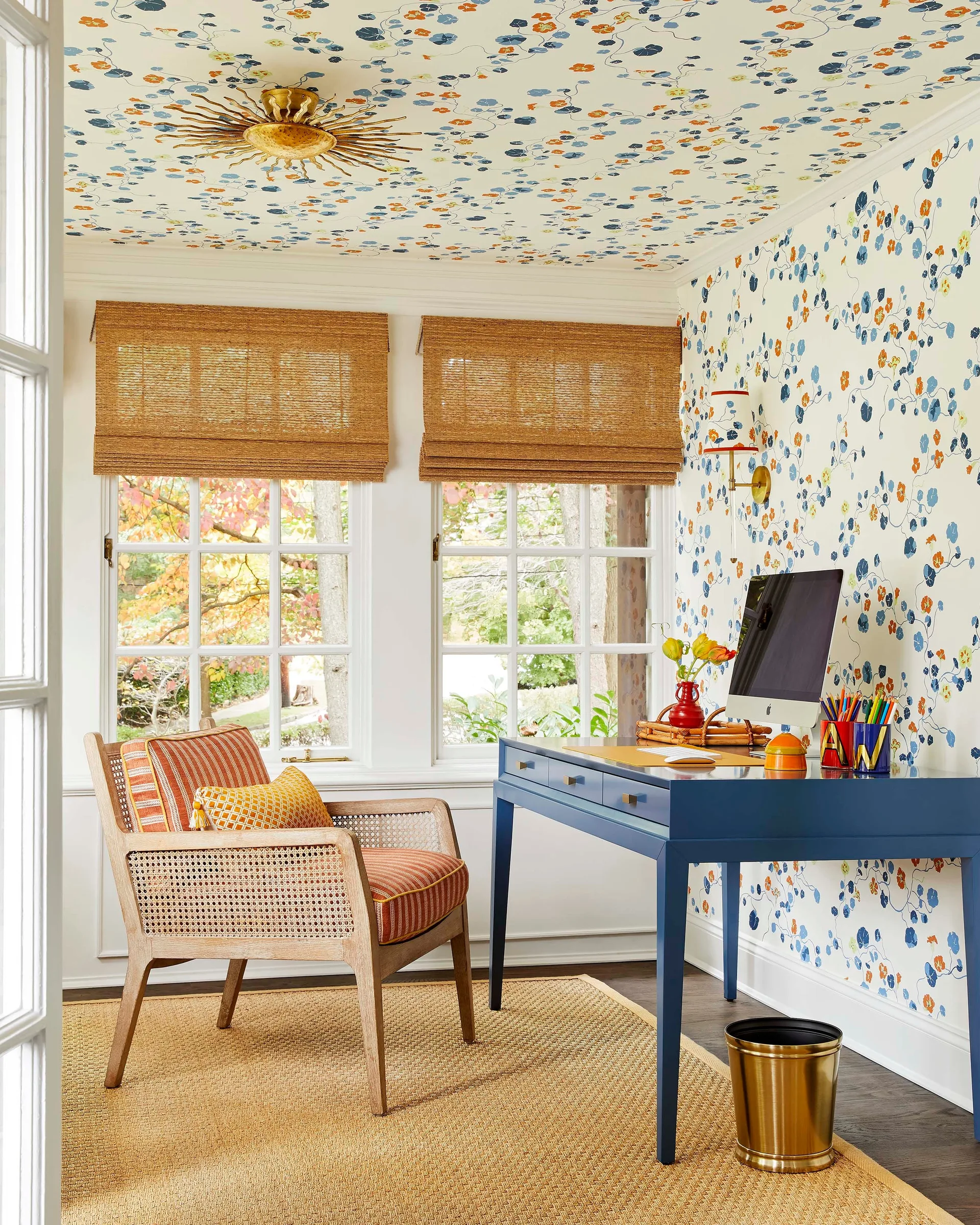 A home office features a blue desk, a cane armchair, and matching floral wallpaper that covers both the walls and the ceiling.