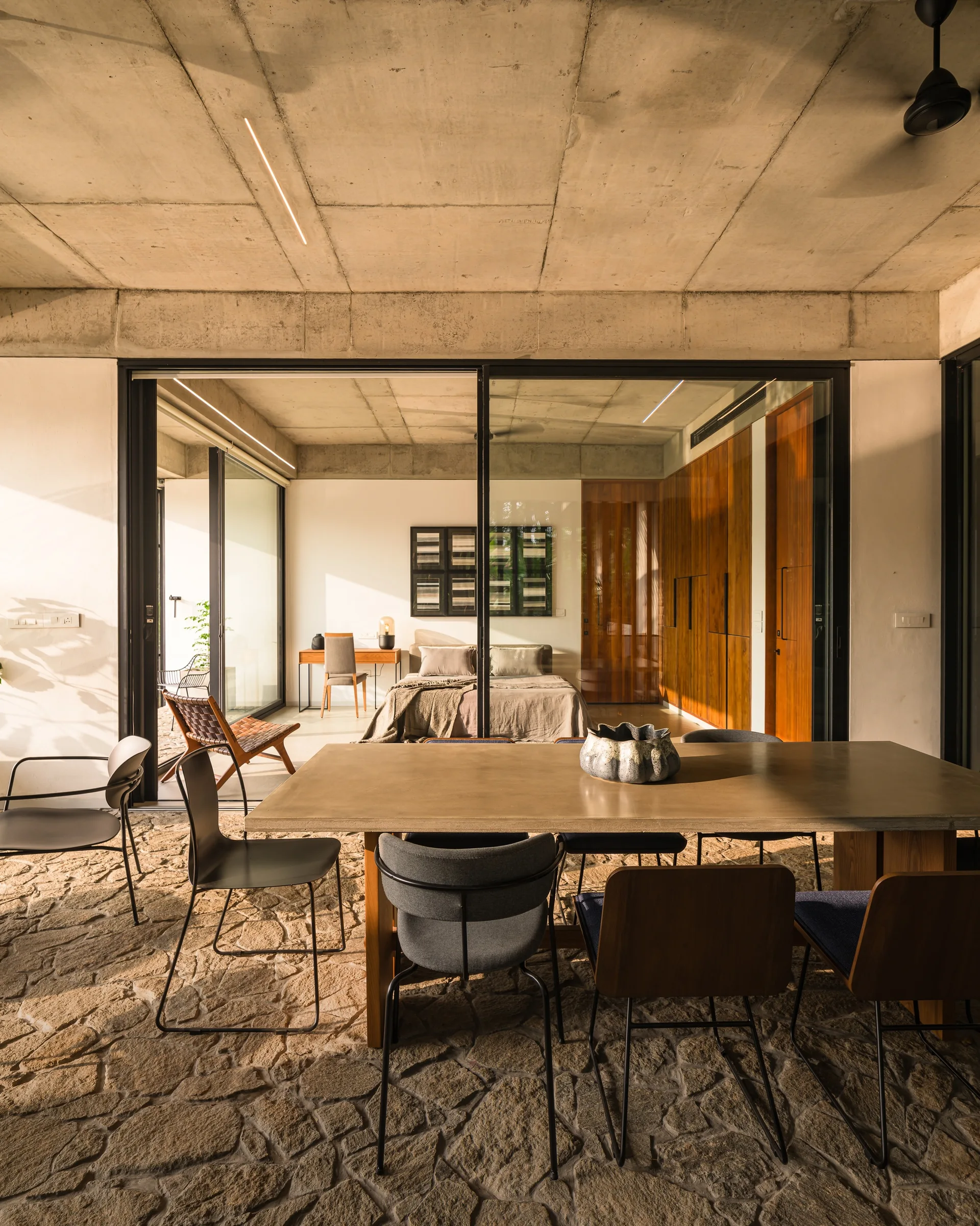 An indoor dining area features a large table on a flagstone floor, with sliding glass doors opening to a bedroom with warm wood cabinetry.