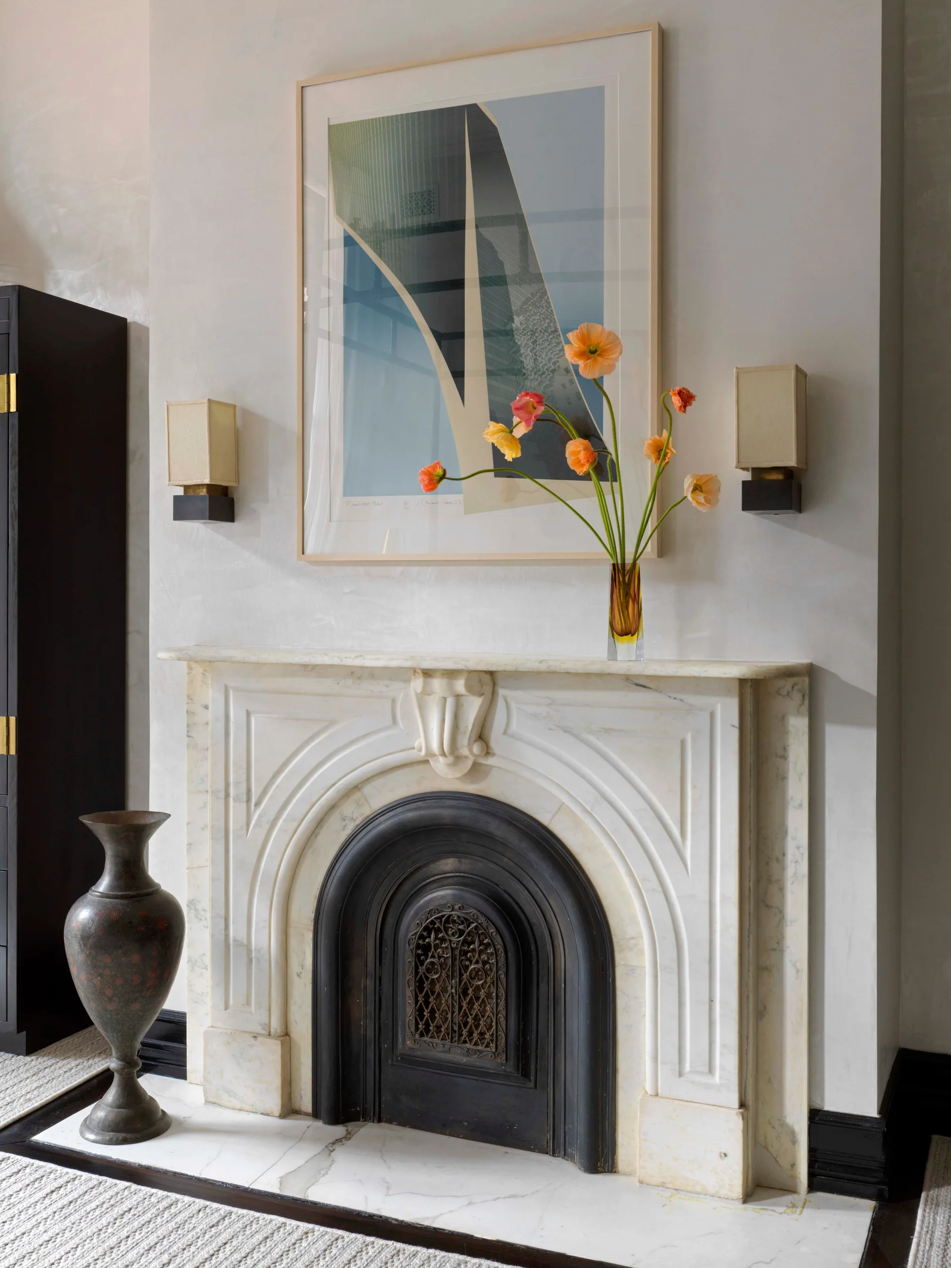 A white marble fireplace with a curved black metal insert features a framed abstract artwork, two rectangular wall sconces, and a vase of orange poppies.