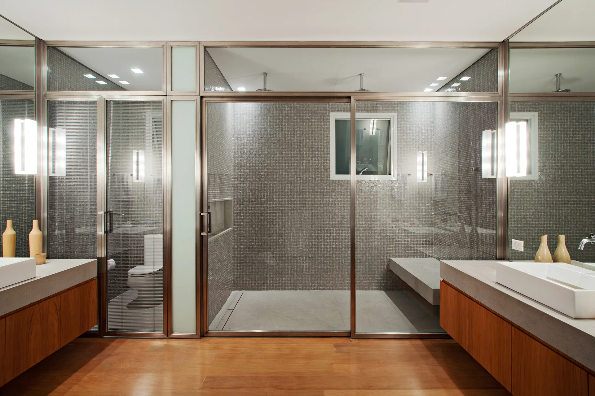 A modern bathroom featuring a large glass-enclosed shower stall with grey mosaic tiles flanked by two wood-toned vanities with white vessel sinks.