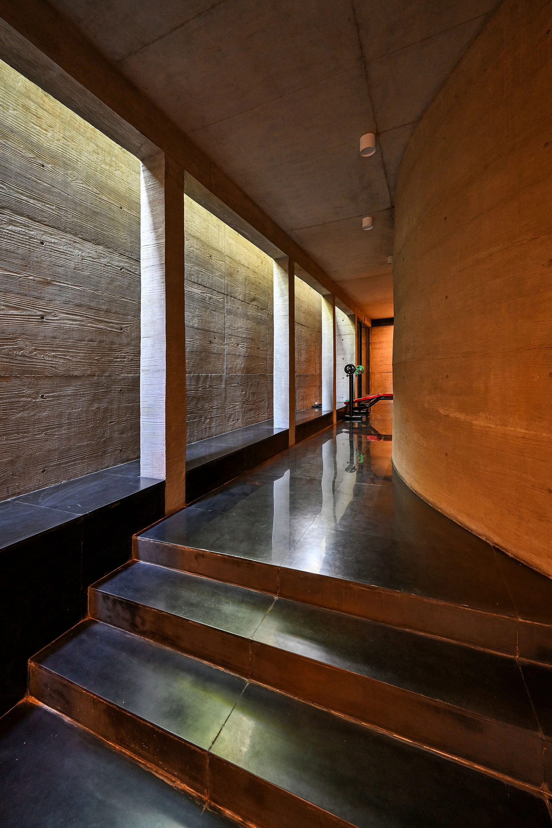 A concrete hallway features board-formed textured walls, a curved wall on one side, and highly reflective dark polished steps and flooring.