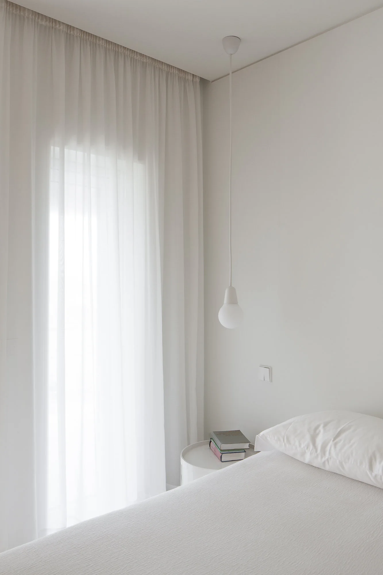 A minimalist white bedroom corner features a bed with white bedding, a round side table, and a hanging white globe pendant light.